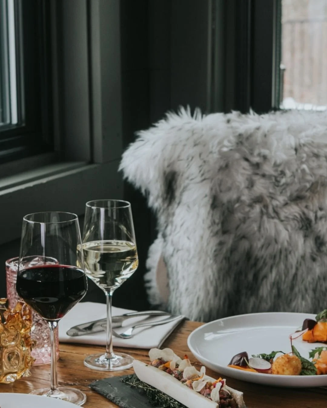 A cozy table setting with two glasses of wine, one red and one white, on a wooden table with assorted food dishes, near a window with a view of a furry blanket or pillow