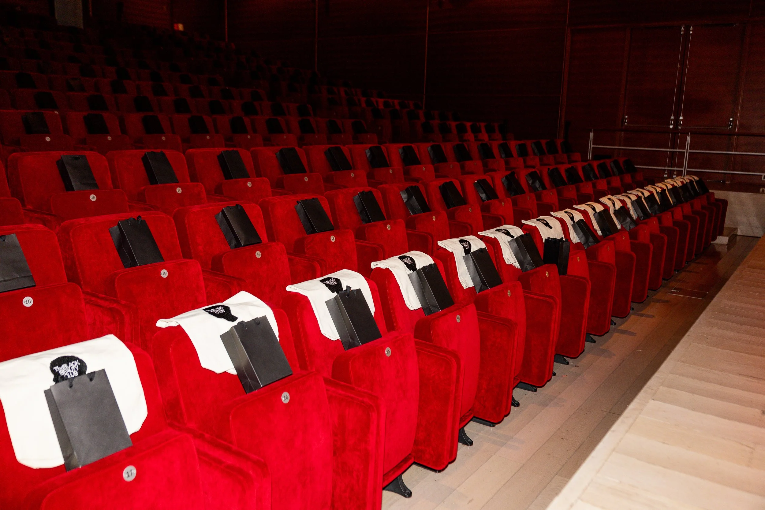 Empty red theater seats with black gift bags and white T-shirts placed on some of the seats in an auditorium.