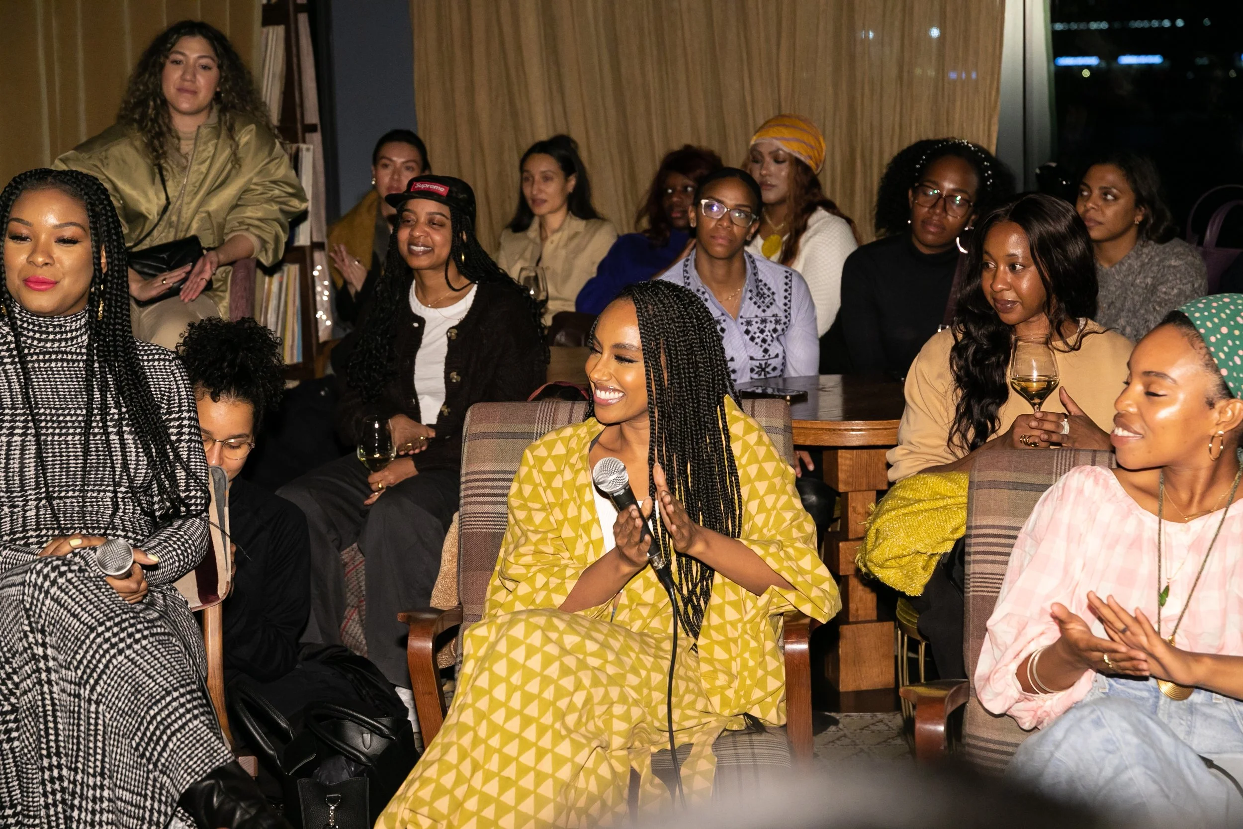 A group of women sitting and standing in a room, some holding drinks, and one woman in yellow with braids holding a microphone, smiling and speaking.
