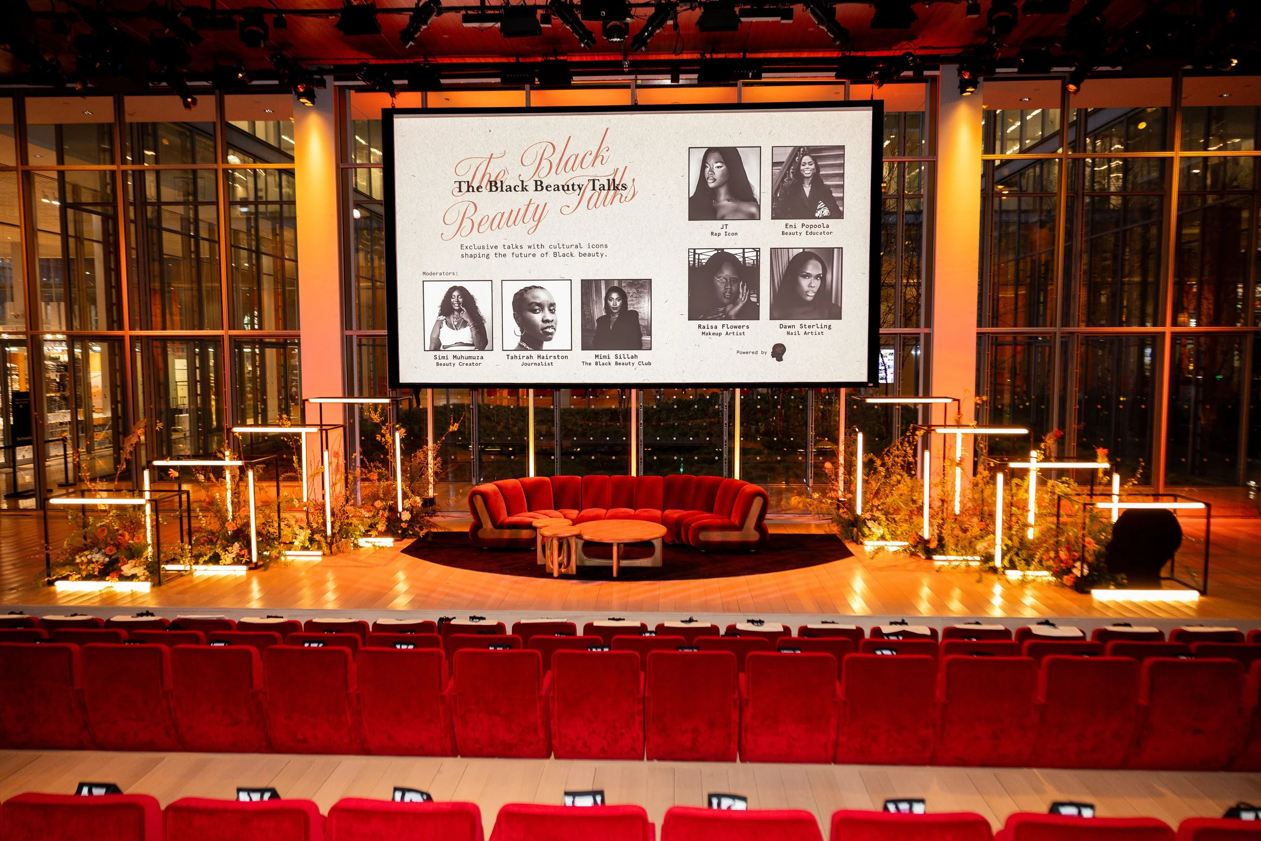A stage set up for a talk called "The Black Beauty Talks" with a large screen displaying profiles of speakers. The stage features a red velvet sofa, small coffee tables, and floral decorations. Rows of red theater seats face the stage in an auditorium.