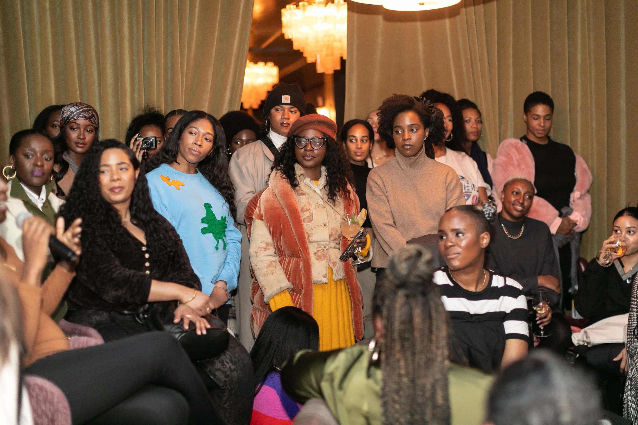 A group of women gathered indoors, some standing and some seated, at a social event with warm lighting and curtains in the background.
