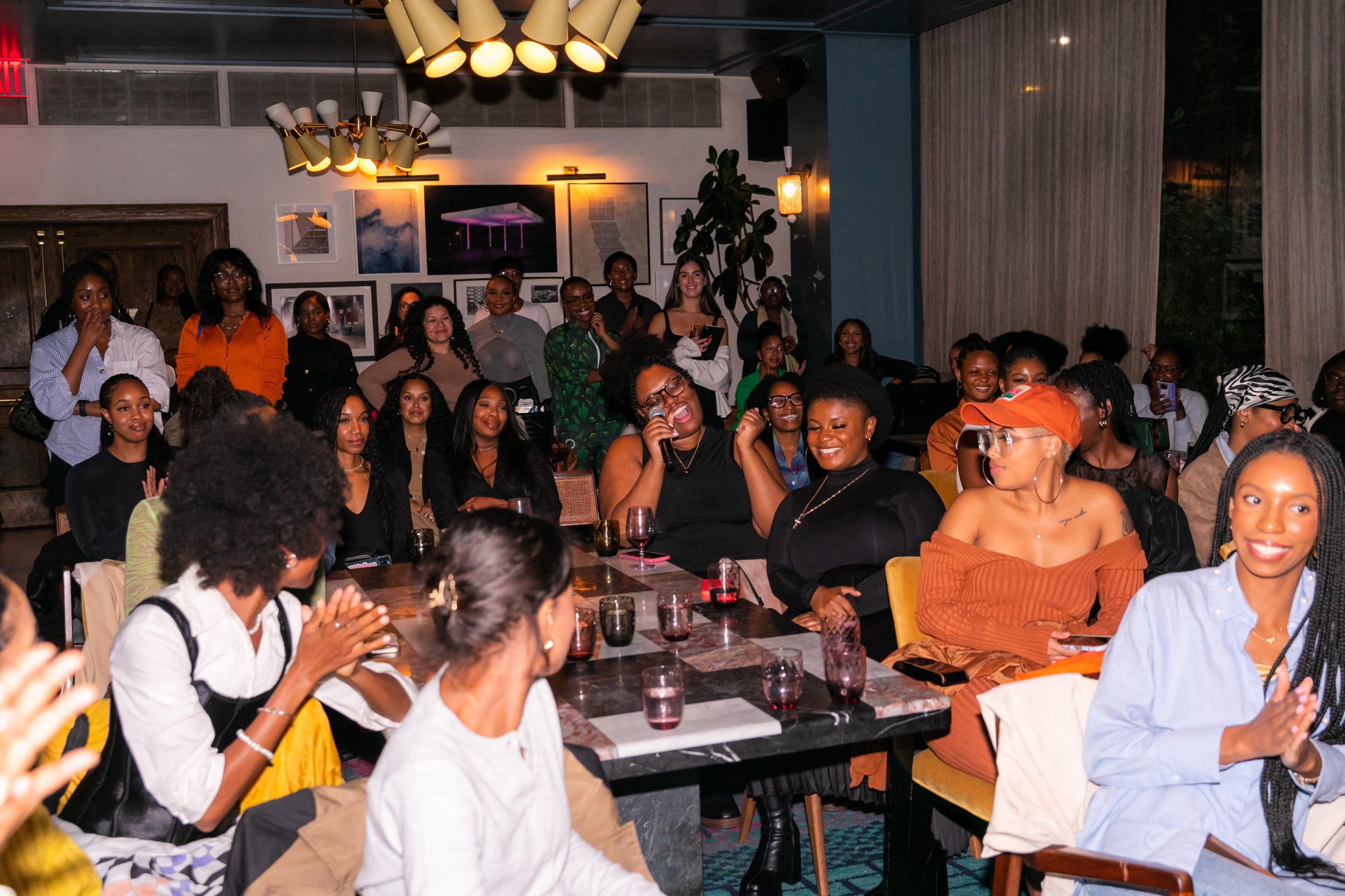 A diverse group of women are gathered in a well-lit room, sitting and standing around tables, engaging and smiling at an event or gathering.