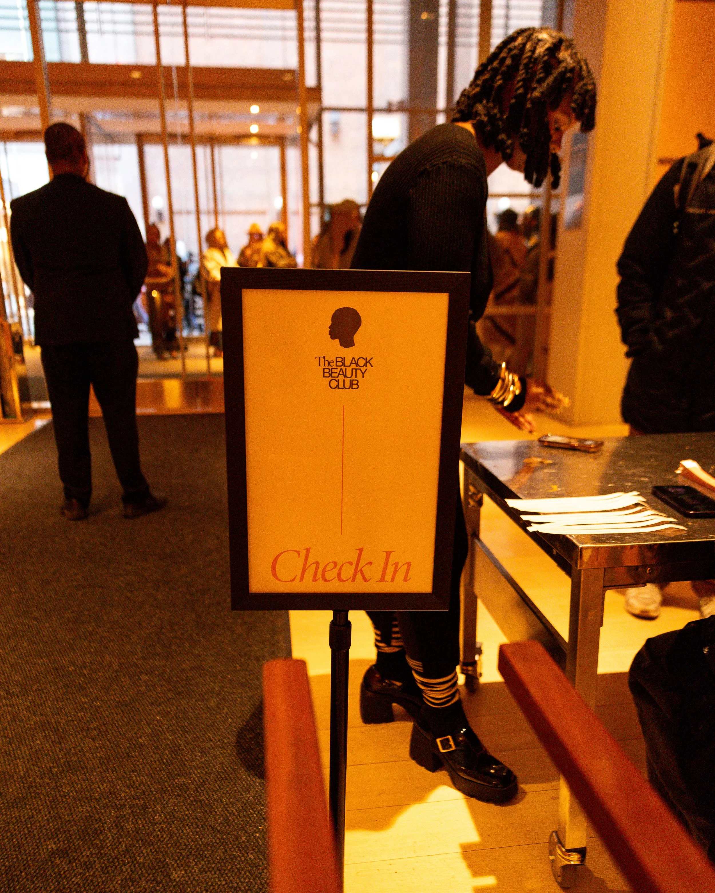 A woman checking in at a reception desk with a sign that reads "Check In" for The Black Beauty Club.