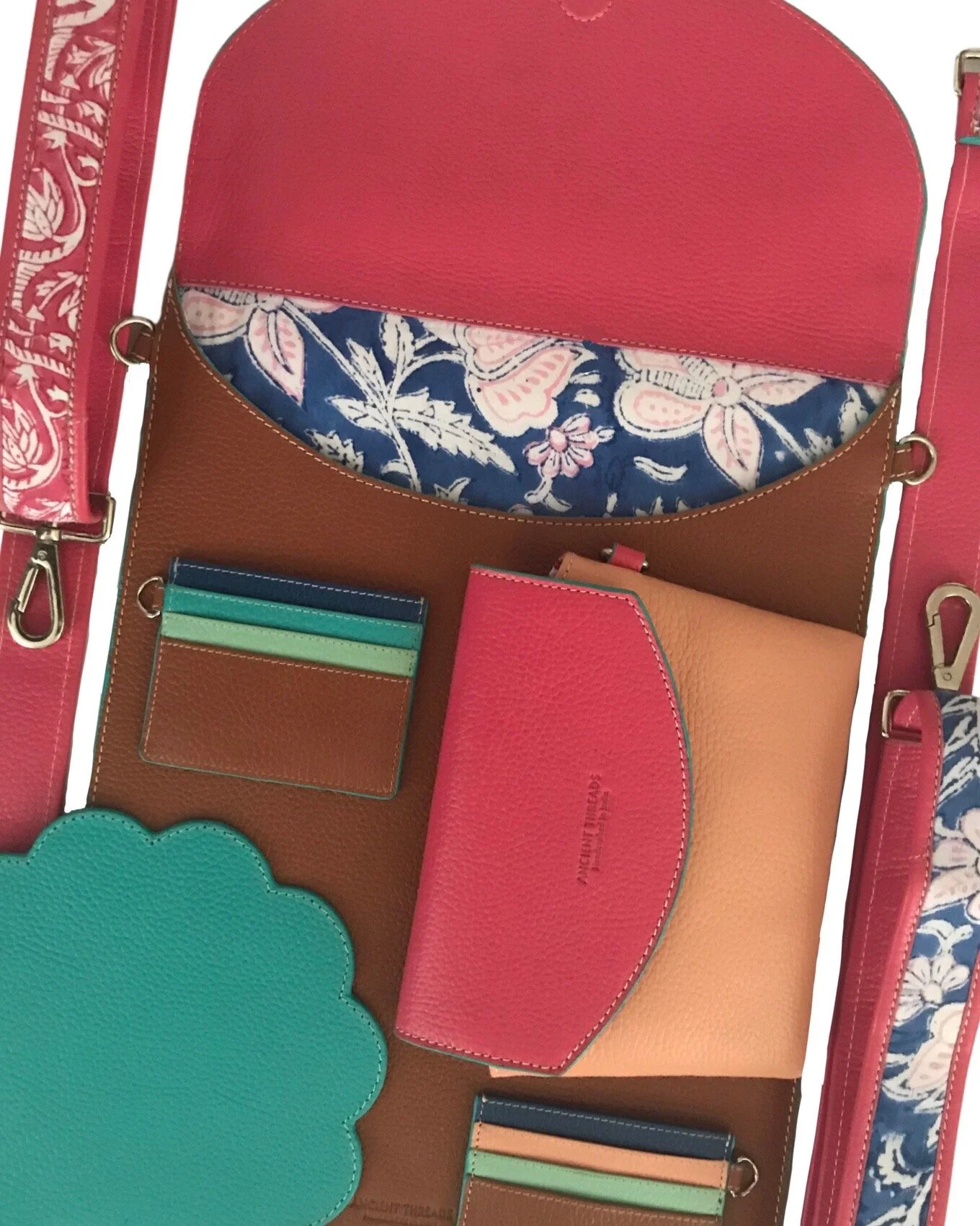Open colorful leather handbag with pink, tan, and blue accents, containing a matching pink wallet, cardholder, and various patterned fabric items, with additional straps nearby.