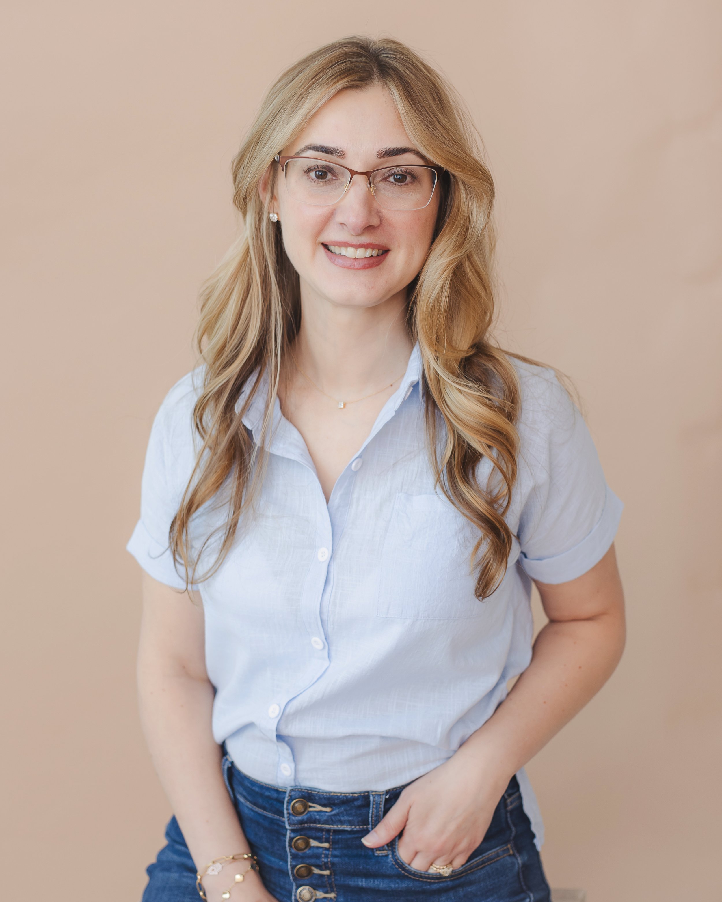 Close-up of an attractive smiling woman with long, wavy blonde hair, glasses, earrings, a light blue button-up shirt, and blue jeans.