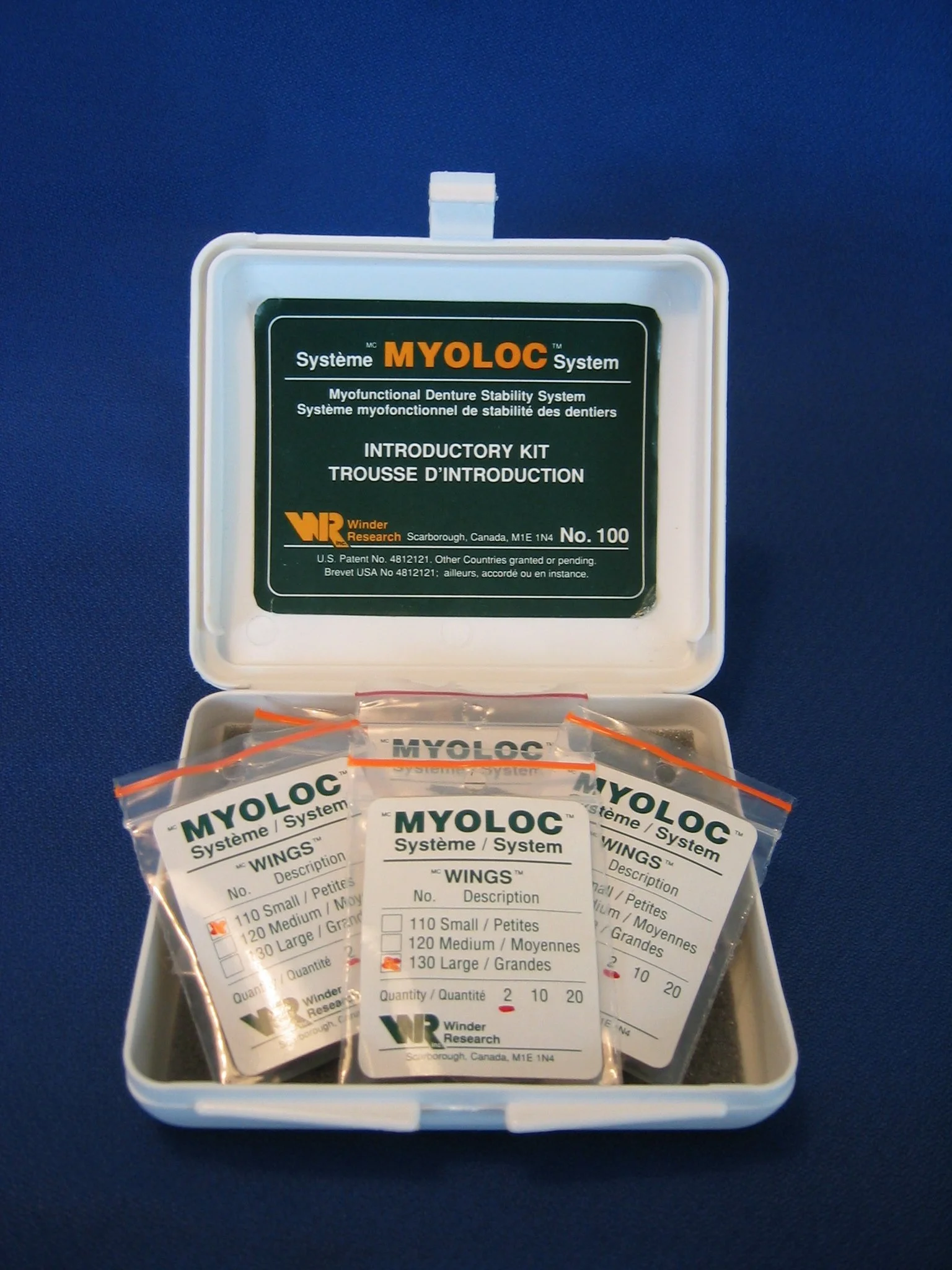 Open plastic box containing small packages labeled MYLOC WINGS for a denture stability system, with a green and white label on the inside of the lid explaining the product.