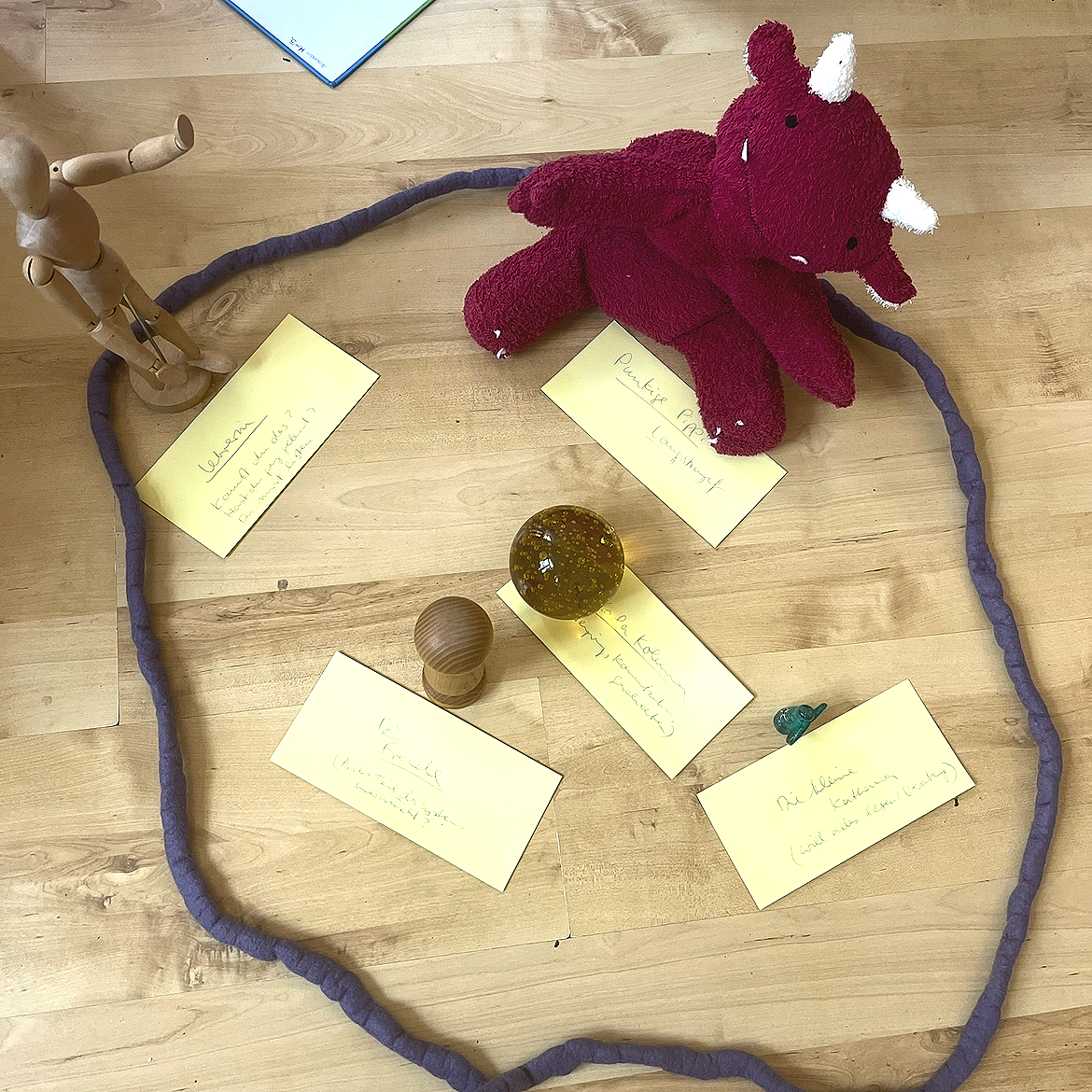 Stofftier-Rhinozeros, eine Puppe, eine Holzpuppe, eine gelbe Glaskugel, eine kleine grüne Figur, mehrere gelbe Notizzettel mit handgeschriebenen Texten, auf einem Holztisch.
