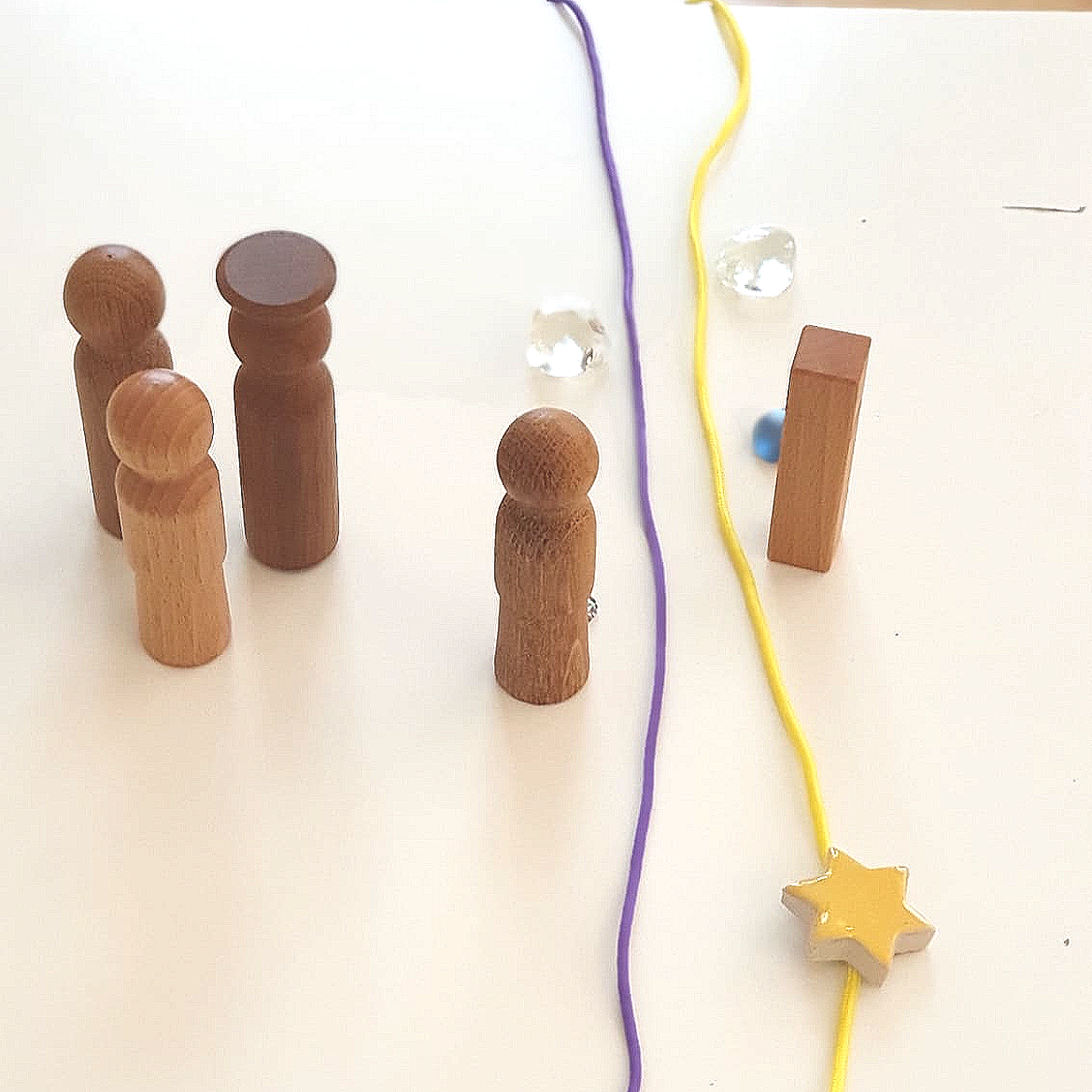 Holzfiguren, bunte Schnüre, Glassteine, Holzklötzchen, eine gelbe Schnur mit einer Sternfigur, auf einem weißen Tisch.