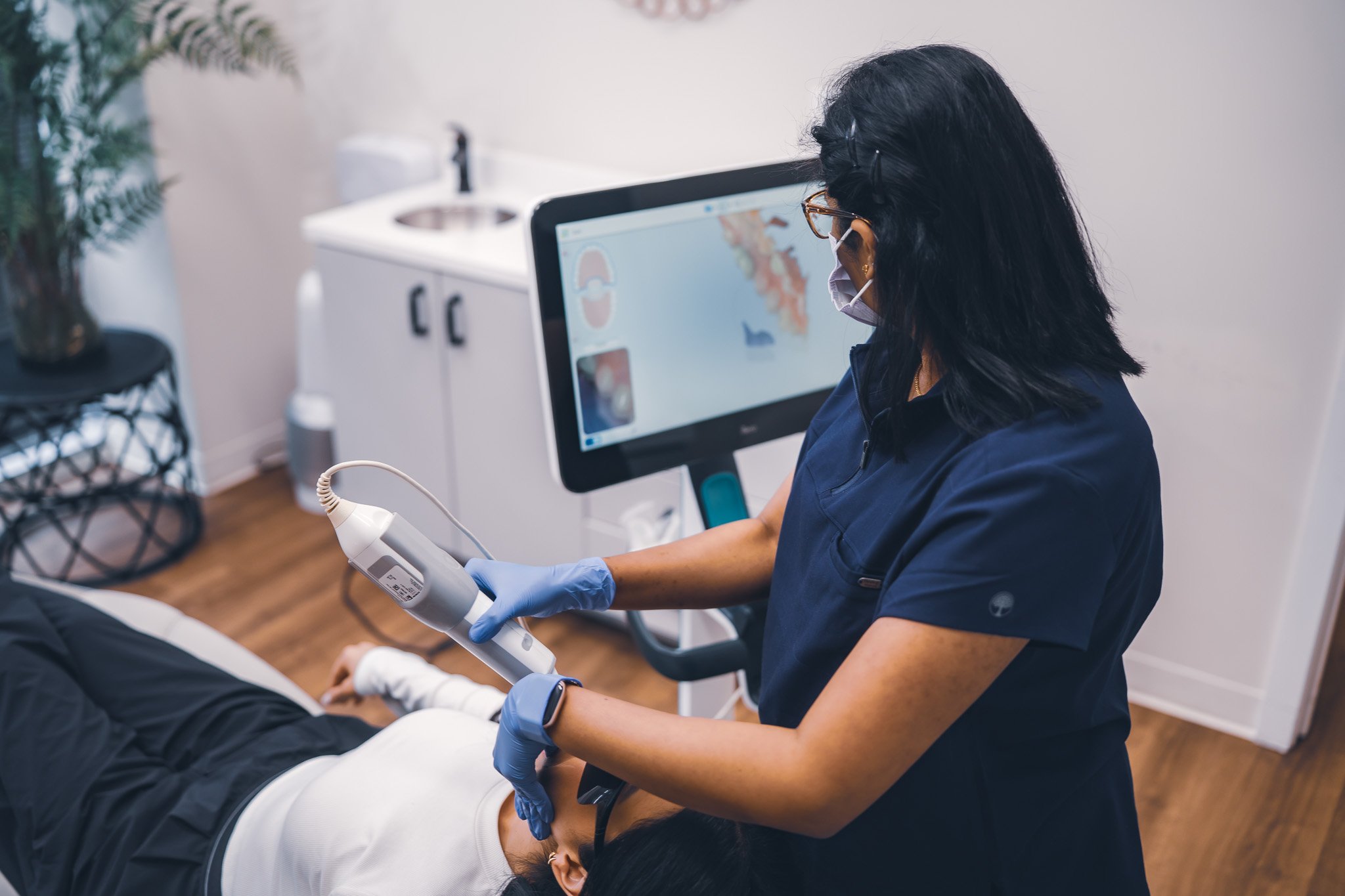 Medical professional wearing a face mask and gloves performing a procedure on a patient's arm with a handheld device in a clinical setting.