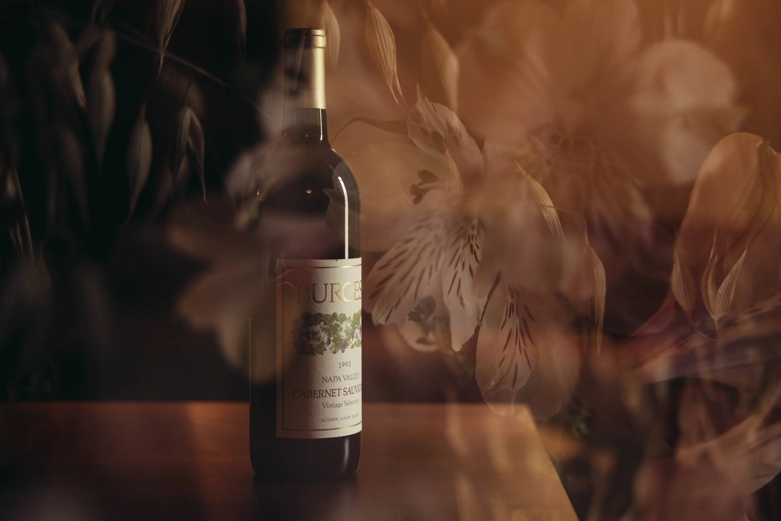 A bottle of wine placed on a wooden surface, surrounded by dried flowers with soft, warm lighting.