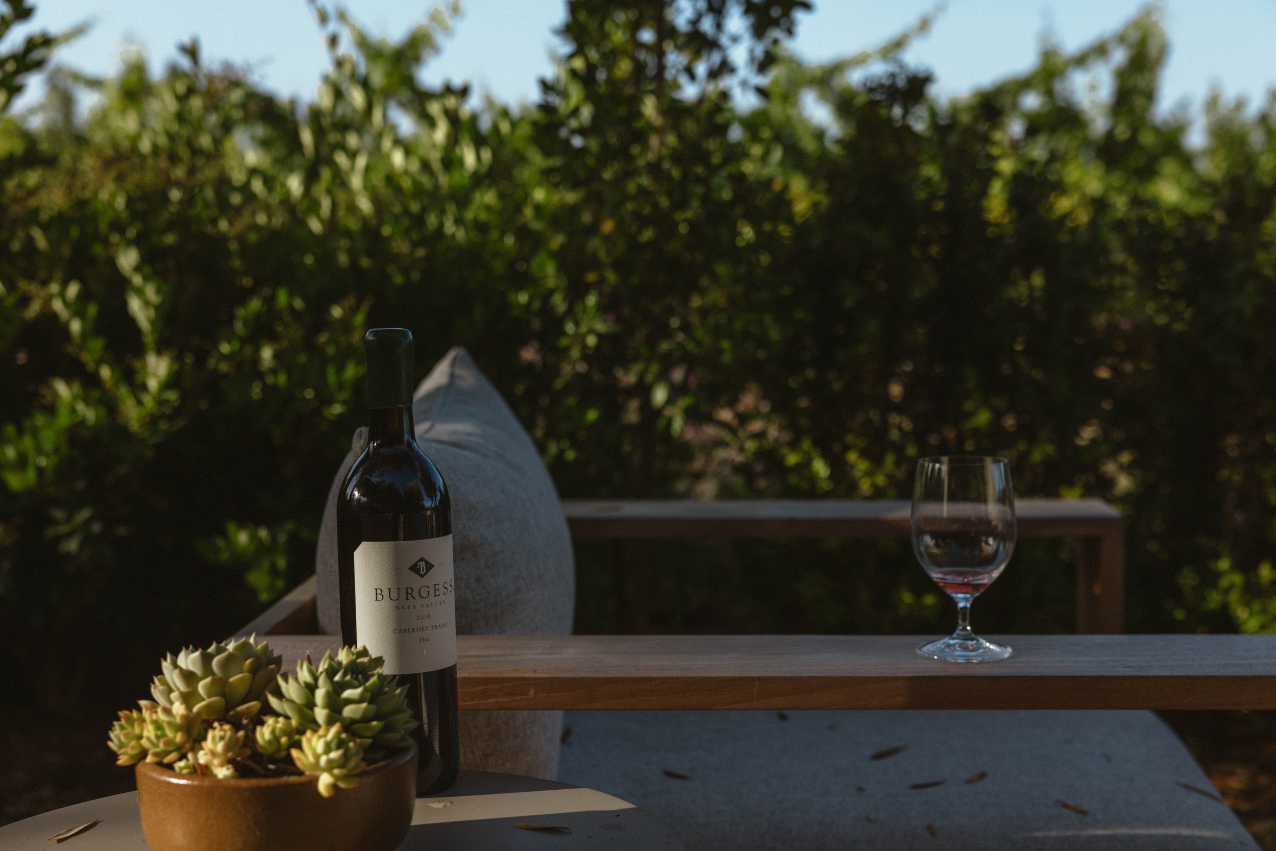 A patio table with a potted succulent, a bottle of Burgess Napa Valley Cabernet Franc wine, an empty wine glass, and a cloth. In the background, a green leafy hedge and a person’s legs are visible, with the person sitting back and enjoying an outdoor