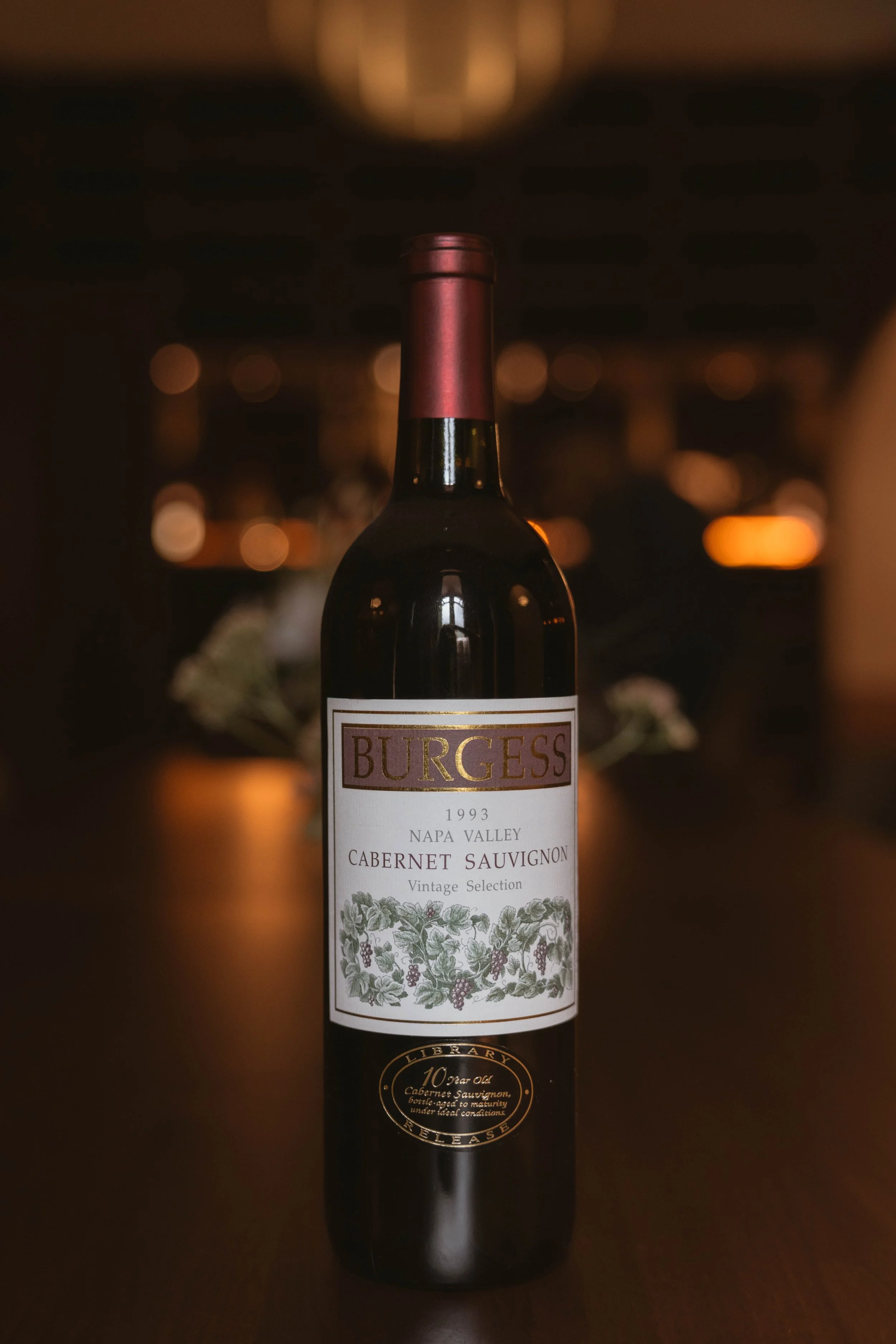 A bottle of Burgess 1993 Napa Valley Cabernet Sauvignon wine on a dark surface with a blurred background of warm lights.