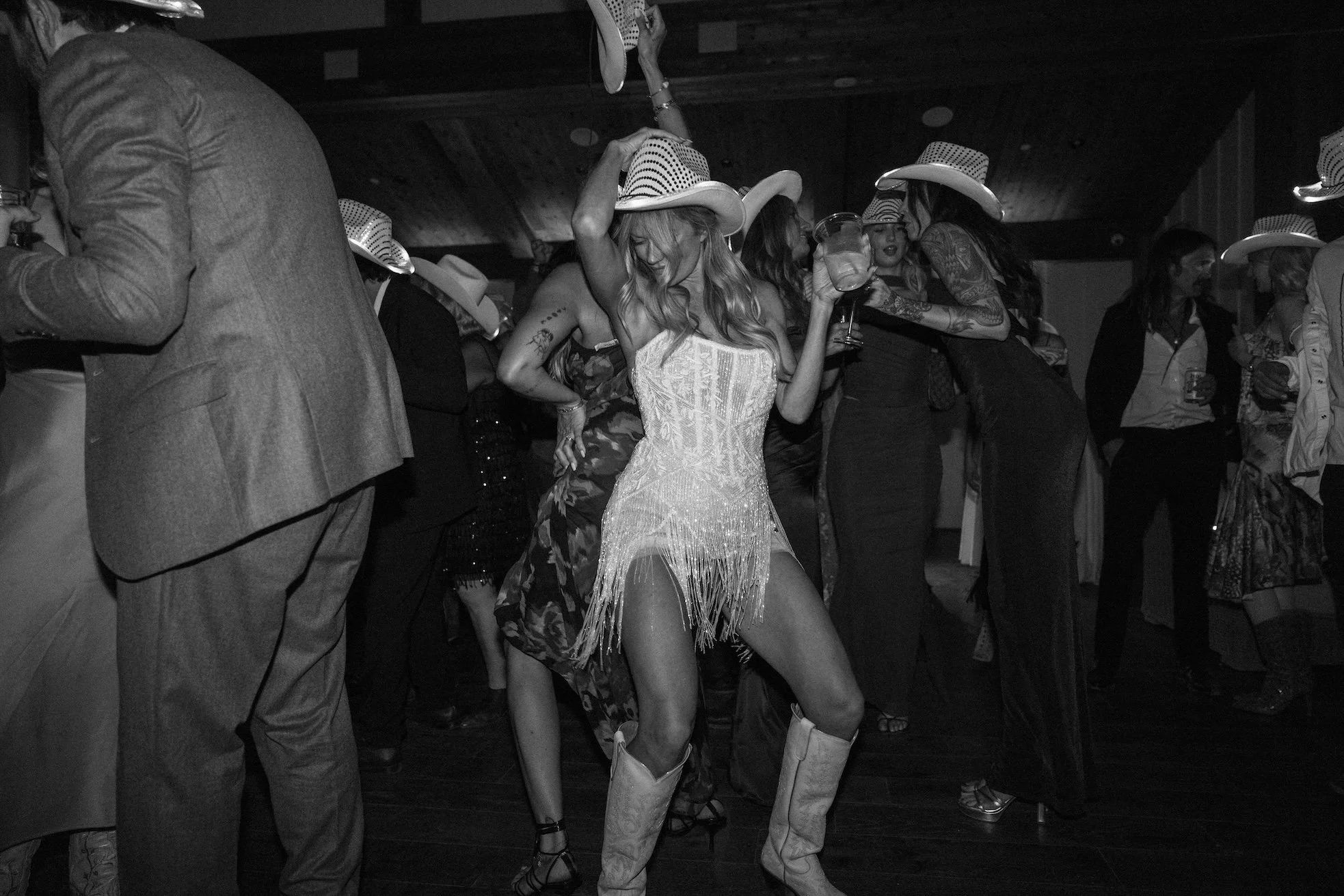 A woman dancing at a party or dance event, wearing a fringed dress and cowboy boots, surrounded by other people also wearing cowboy hats.
