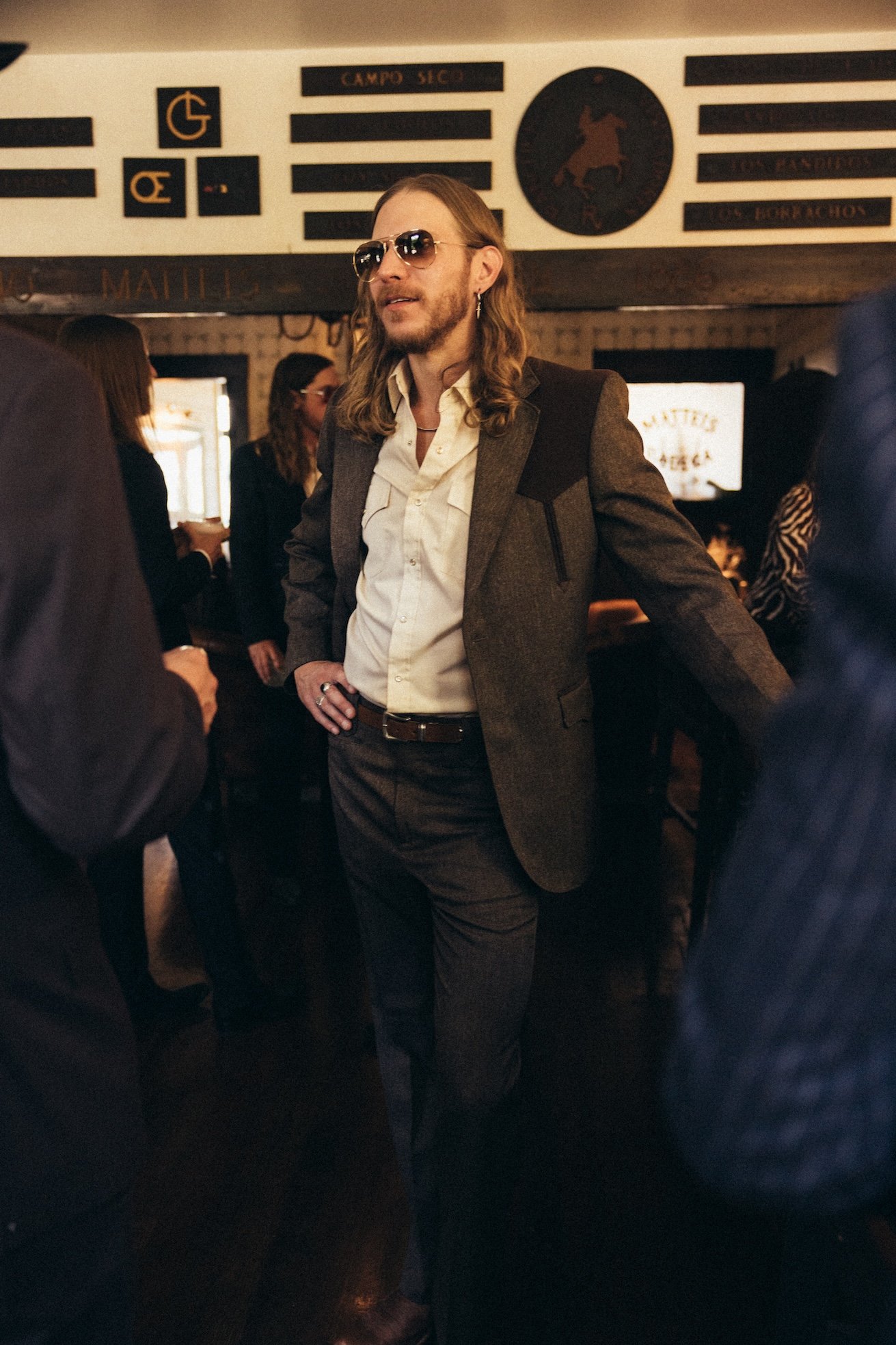 A man with long hair, glasses, and earrings standing in a social gathering or bar, wearing a suit jacket and dress shirt. People are in the background, some looking at their phones, with signs and decorations on the wall behind him.
