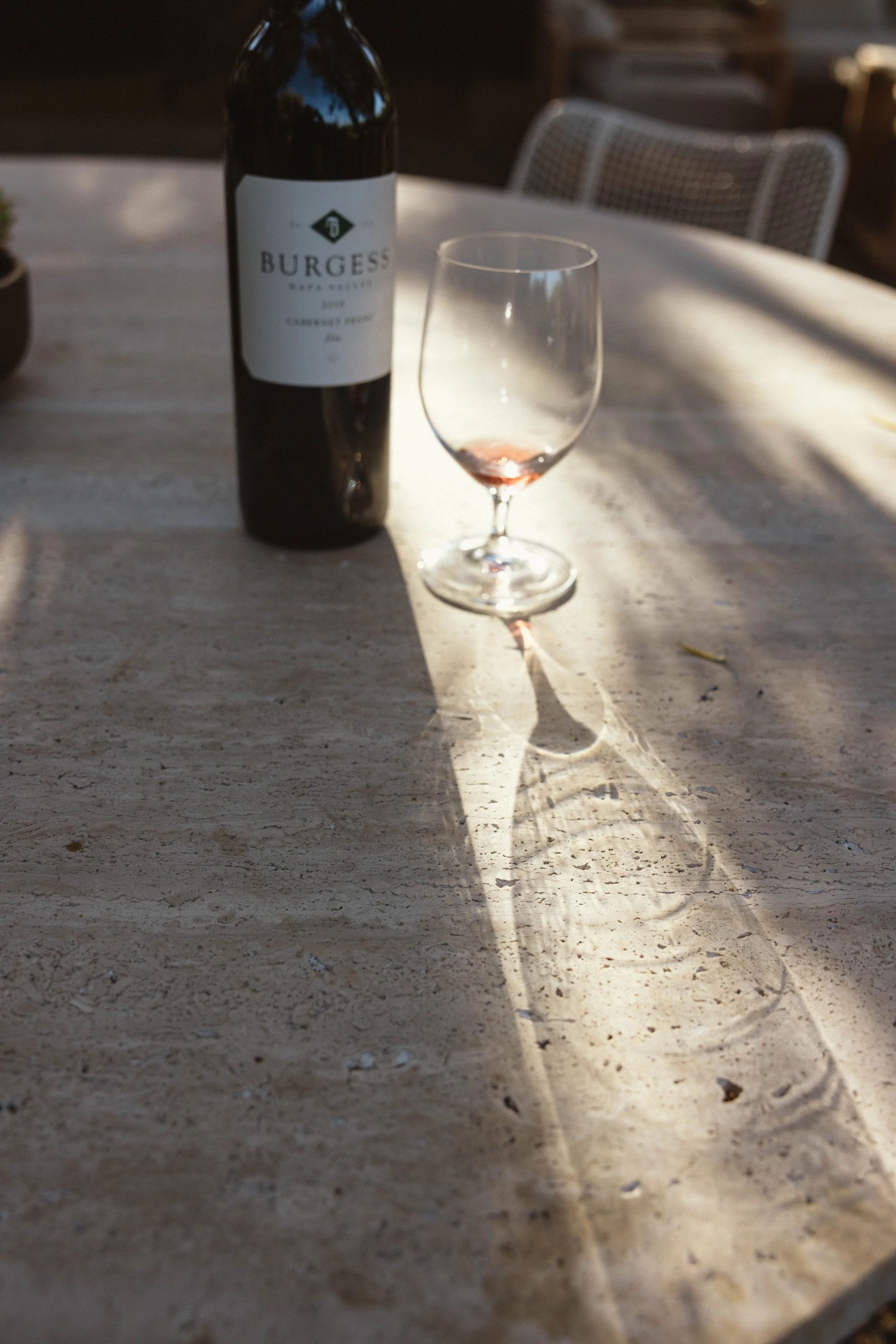 A partially filled wine glass and a bottle of Burgess wine on a light-colored wooden table, with shadows cast by sunlight.