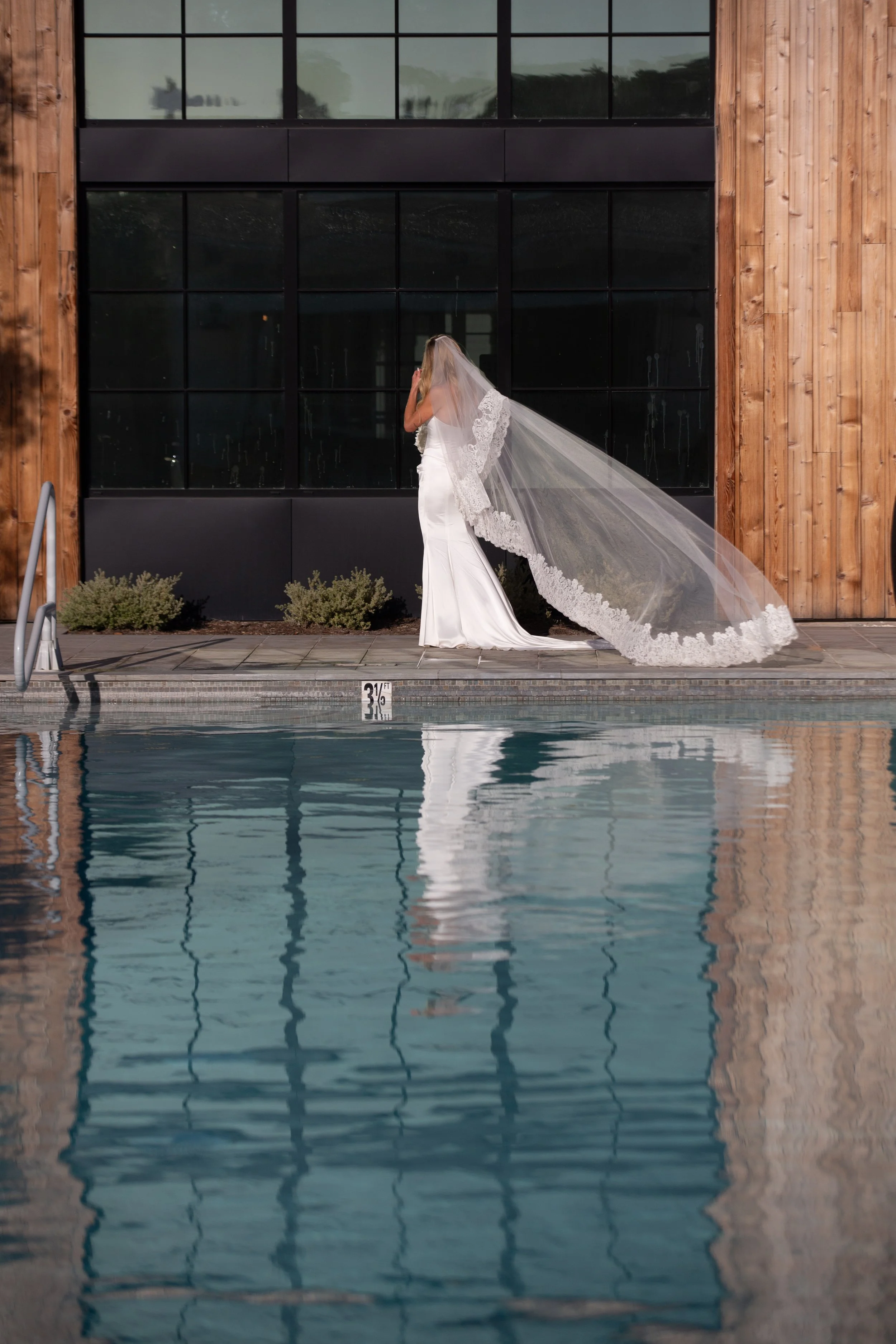 A bride in a white wedding gown with a lace-trimmed veil stands beside a swimming pool during daytime, facing a large window with a reflection of the sky and trees.