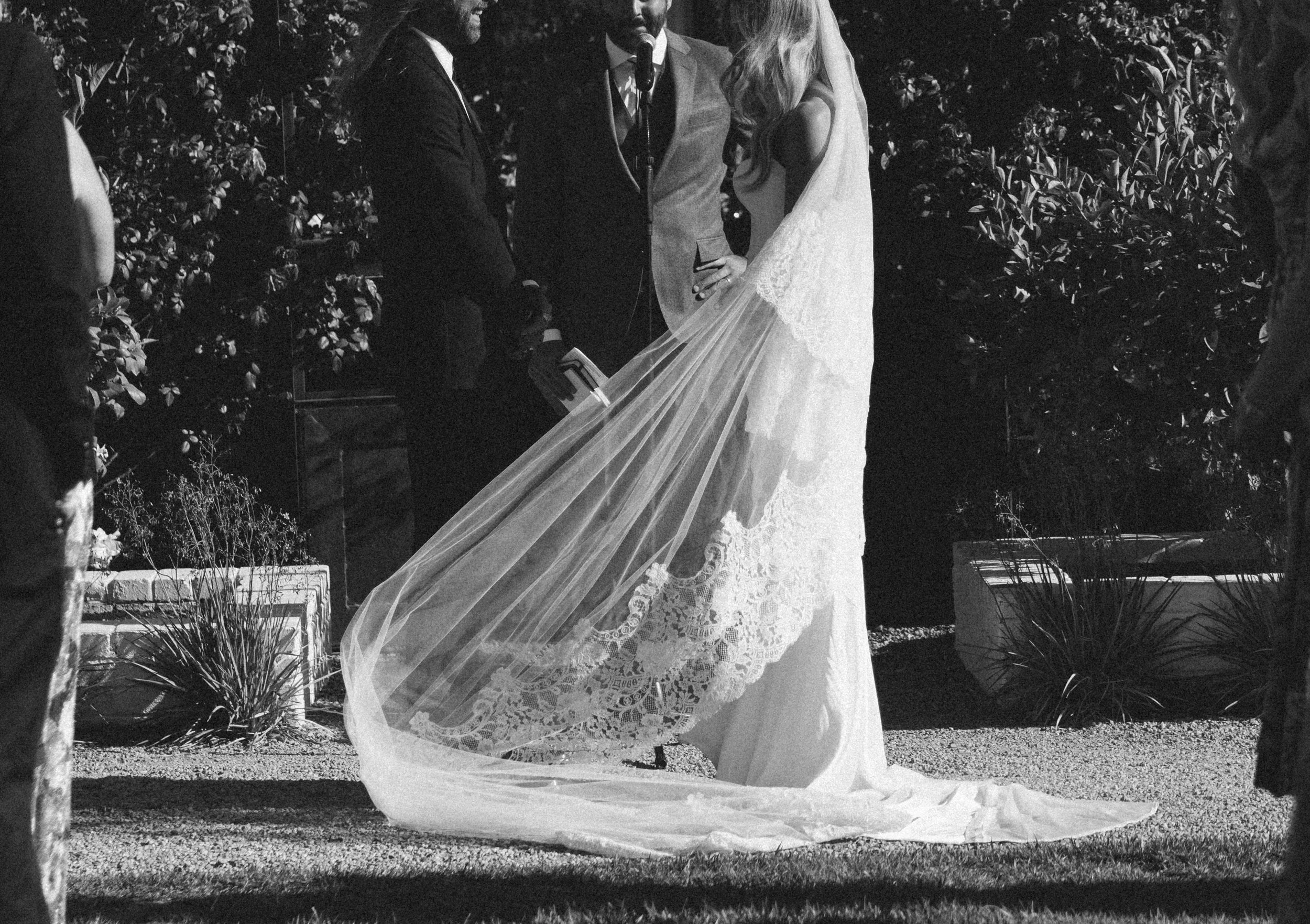 Black and white photo of a bride and groom exchanging vows outdoors, surrounded by trees and bushes at night.