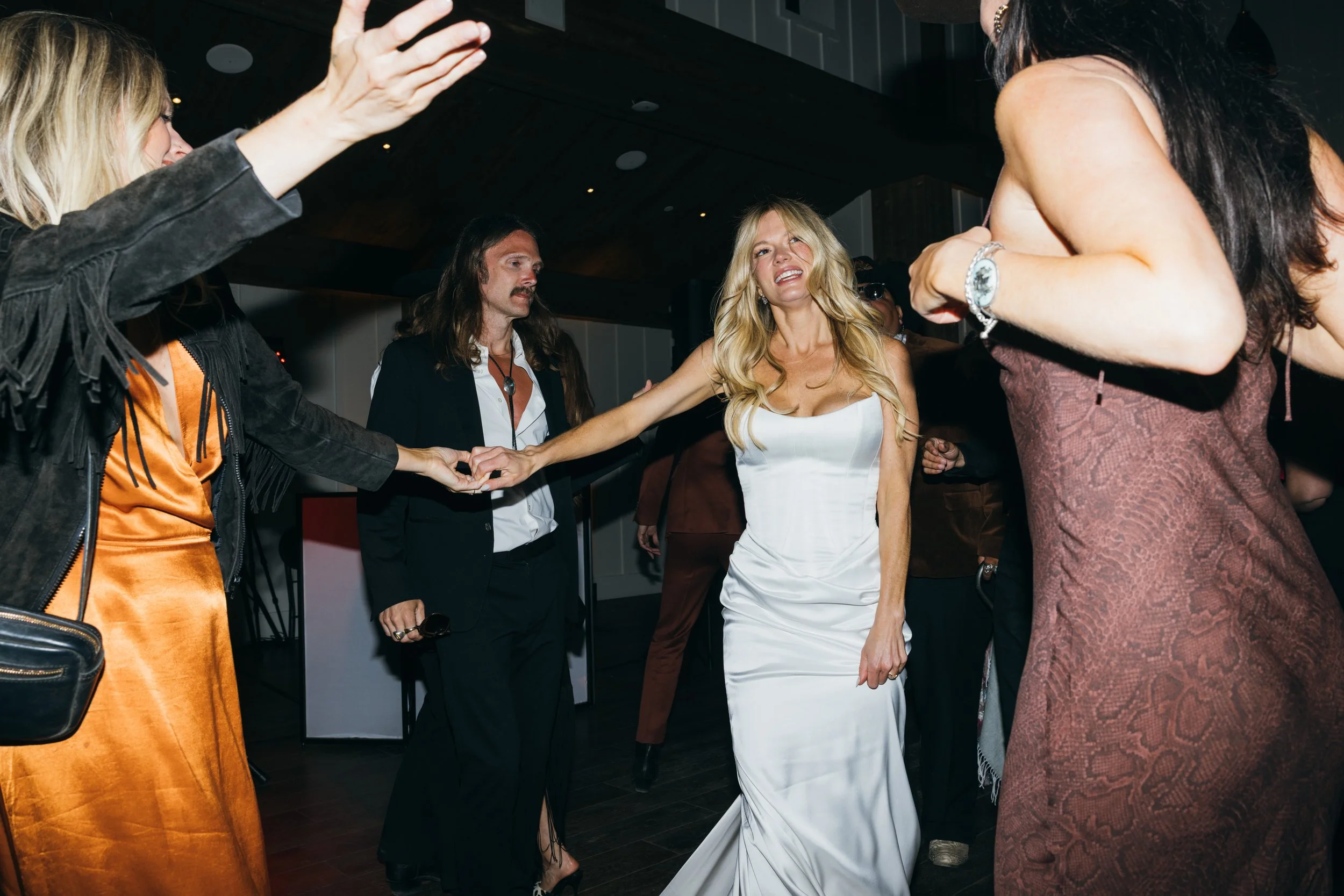 A woman in a white dress dancing with a man in a suit, surrounded by other women in a social setting.
