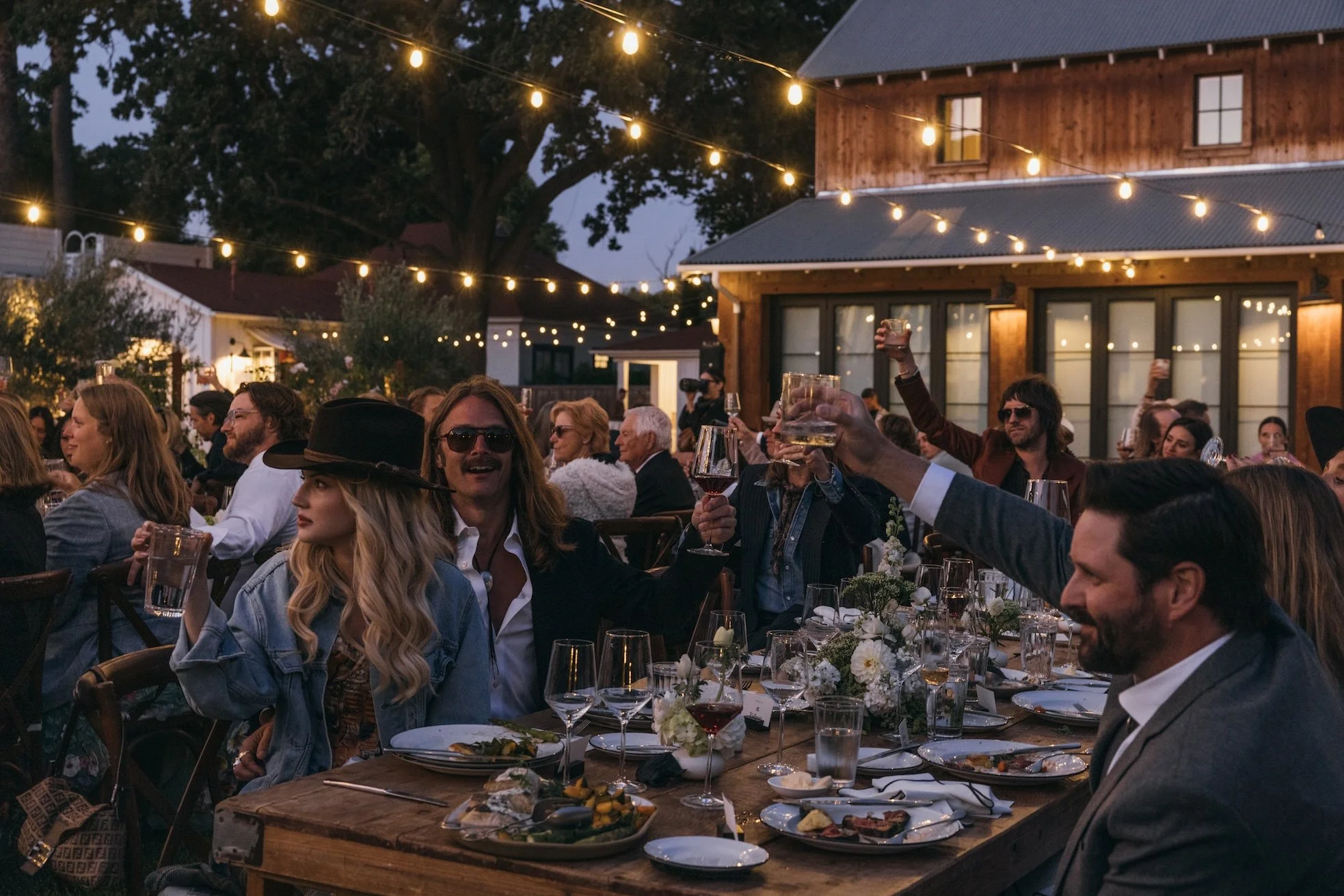 People gathered outside for an evening celebration, sitting at a long table with wine glasses, plates, and centerpieces, with string lights overhead and a rustic wooden building in the background.