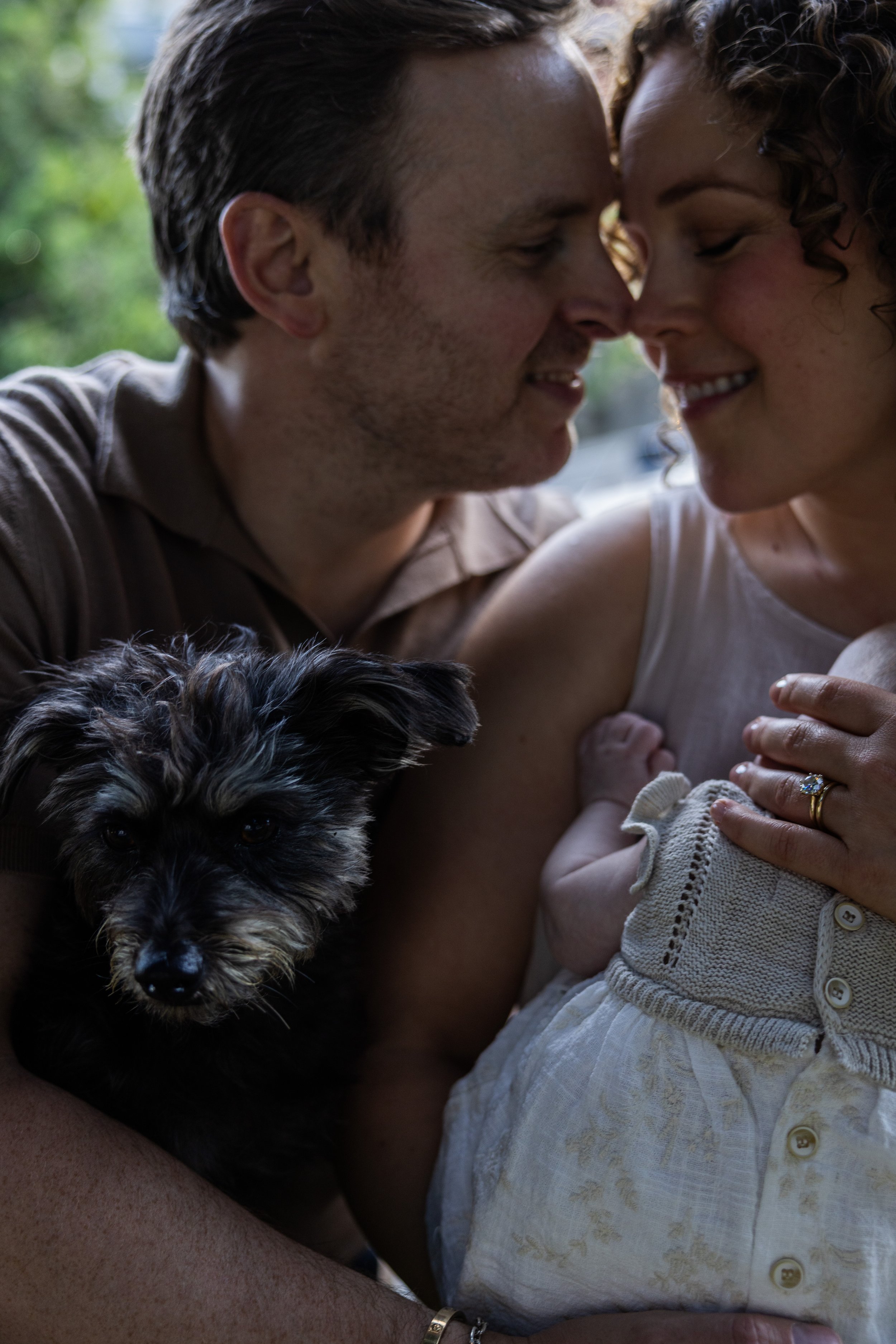 A close-up photo of a couple with their foreheads touching, smiling with closed eyes, holding a young child. The woman has curly hair, and the man has short brown hair. A small black and gray puppy is also in the foreground, looking at the camera. Th