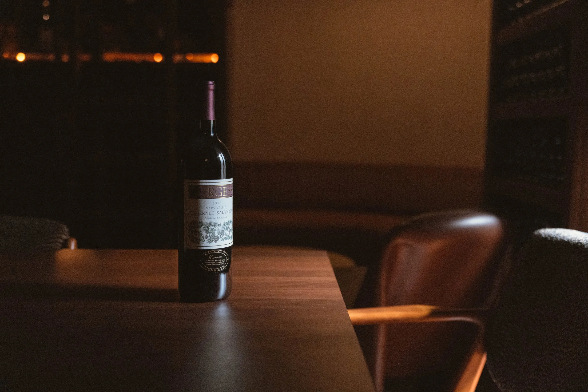 A bottle of red wine on a wooden table in a dimly lit room, with chairs and shelves in the background.