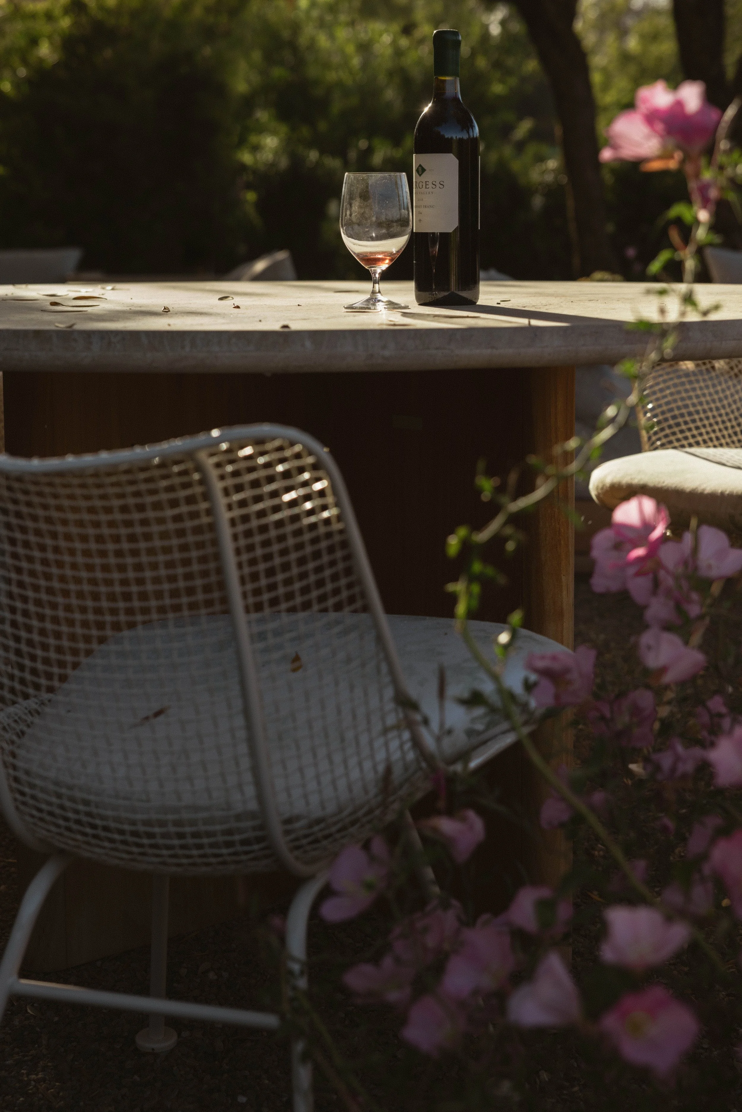 A table outdoors with a bottle of red wine and an empty wine glass, surrounded by chairs and pink flowers, with greenery in the background.