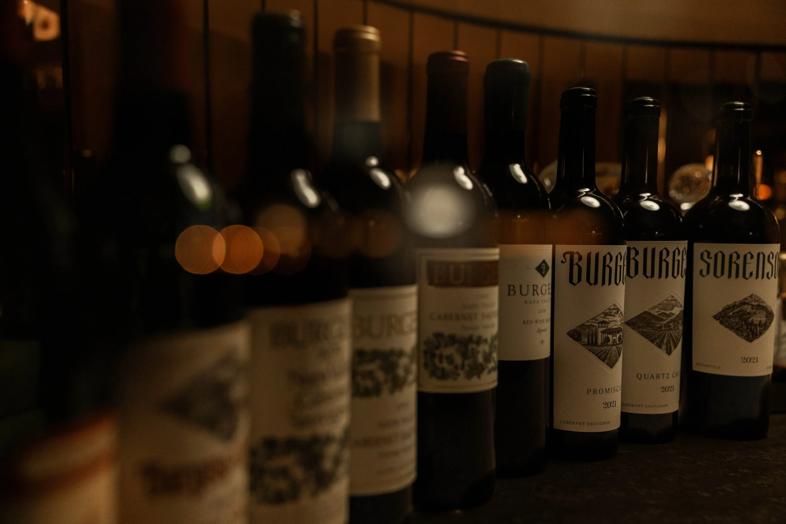 Row of wine bottles on a dark shelf, with some labels visible including 'Burg' and 'Soren'.