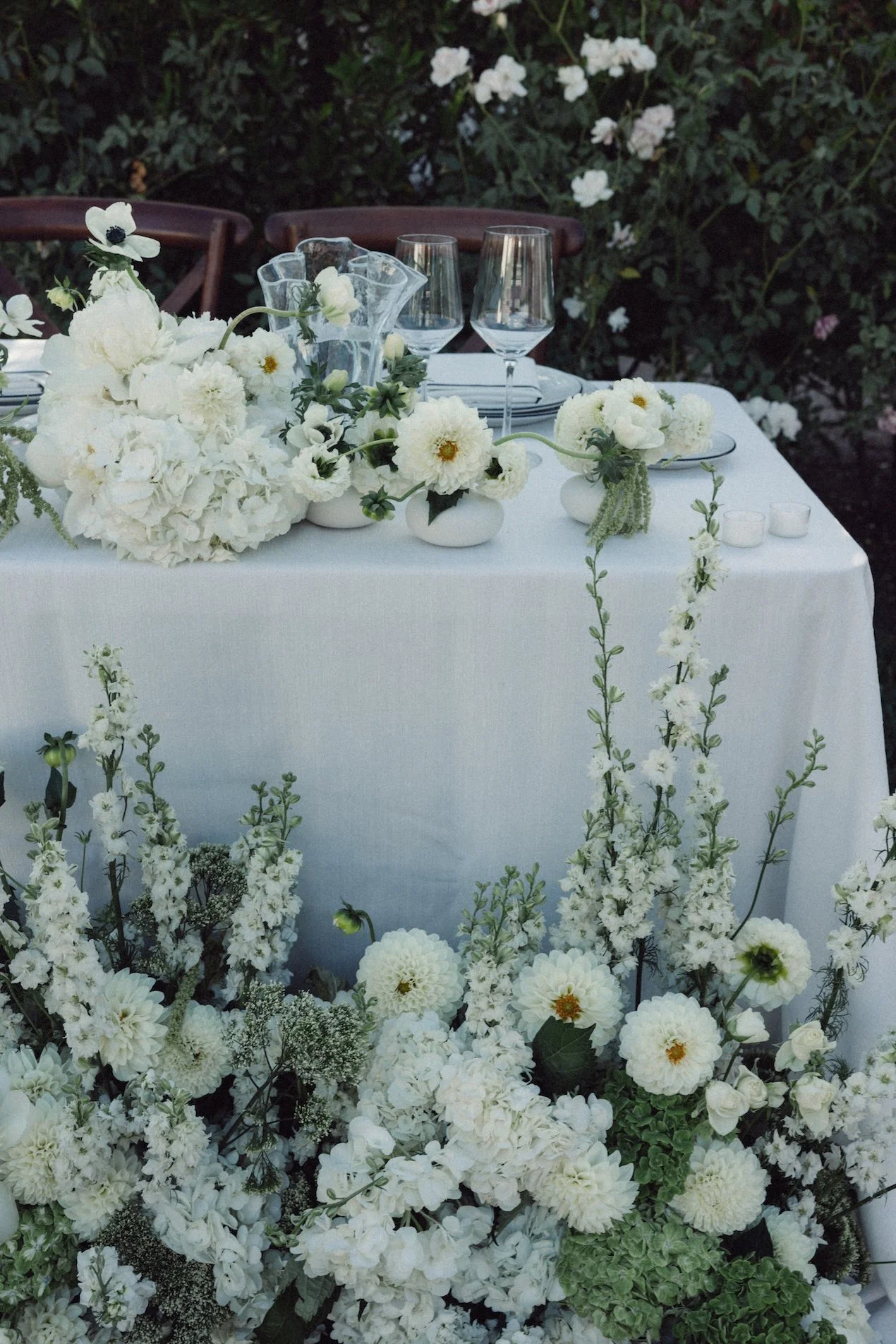 A table decorated with white flowers and glassware set outdoors, surrounded by greenery.