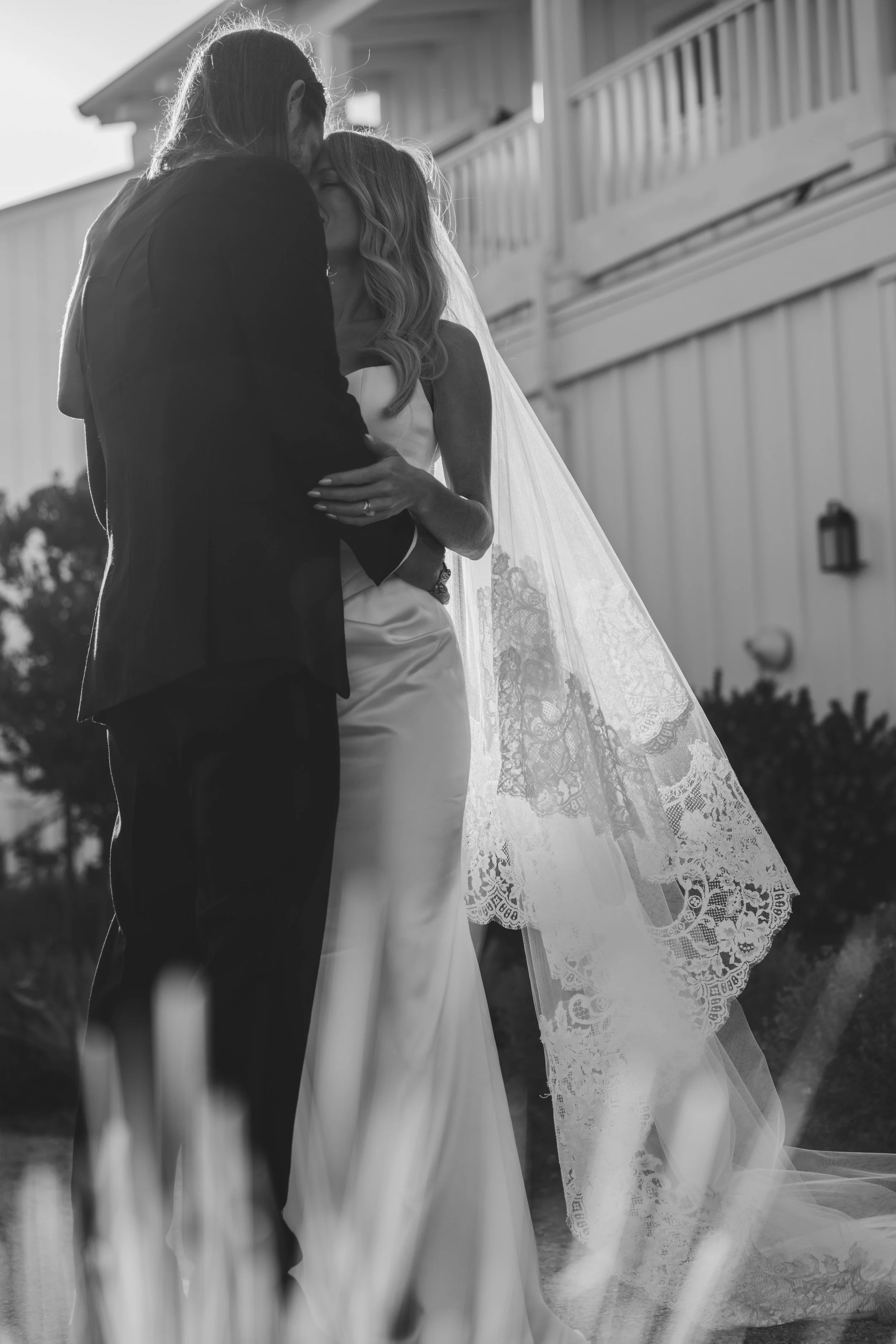 A black and white photo of a couple embracing outdoors, with the woman wearing a wedding dress with lace details and a long veil, and the man in a suit, in front of a building with a balcony.
