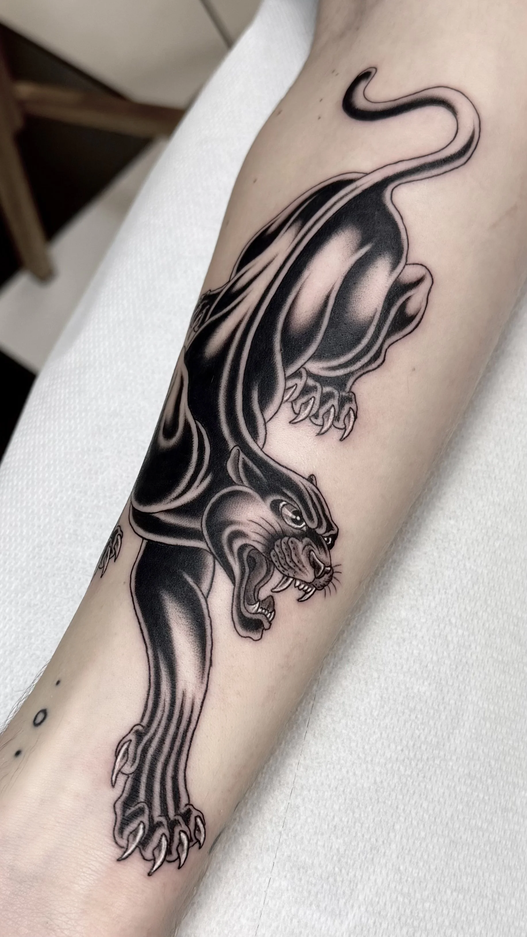 Black and gray tattoo of a panther on person's forearm, showing the animal in a prowling stance with mouth open, claws extended, and tail curled upward.