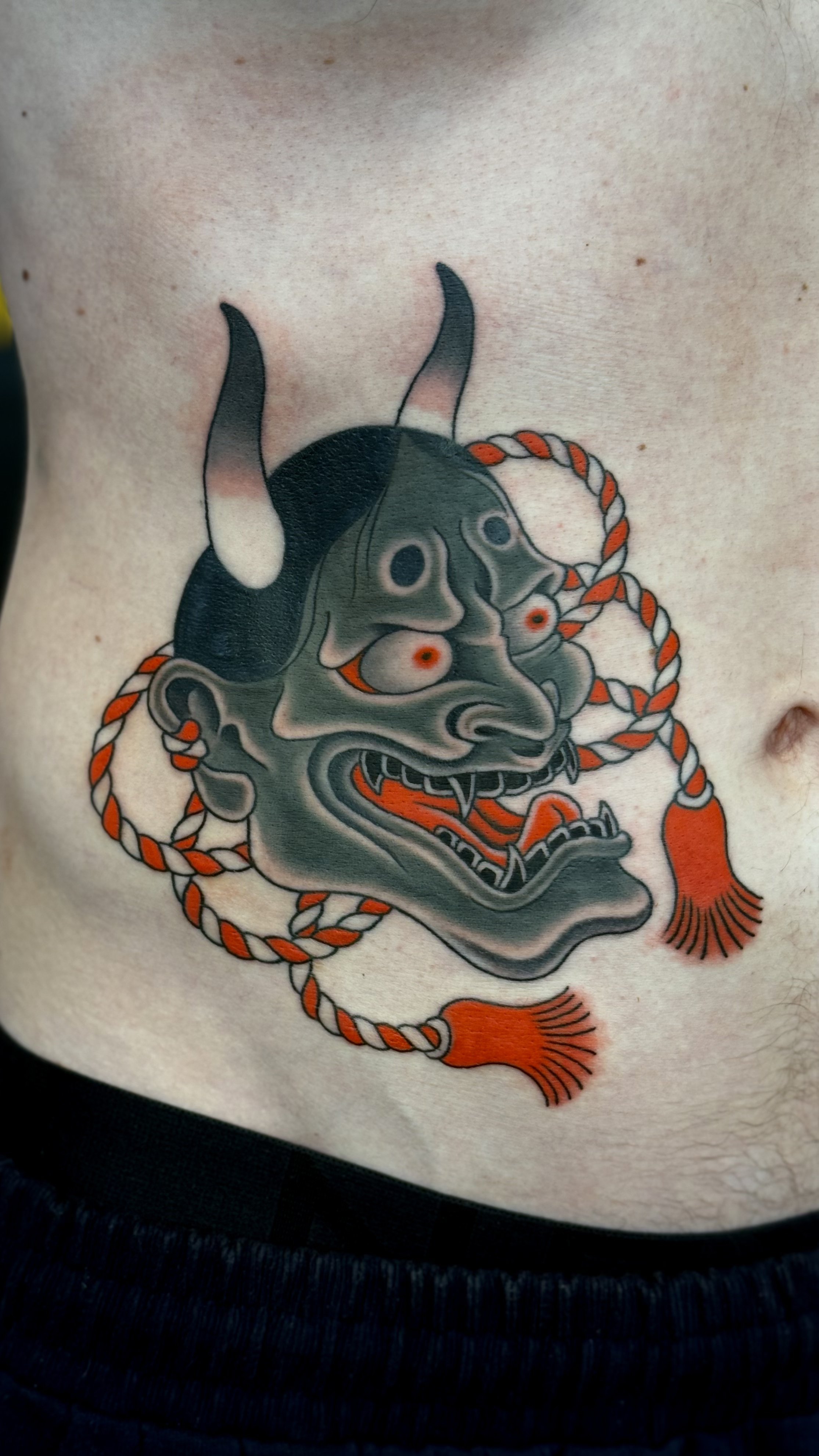 Tattoo of a traditional-style demon or oni mask with horns, twisted rope, and tassels on the person's side torso.