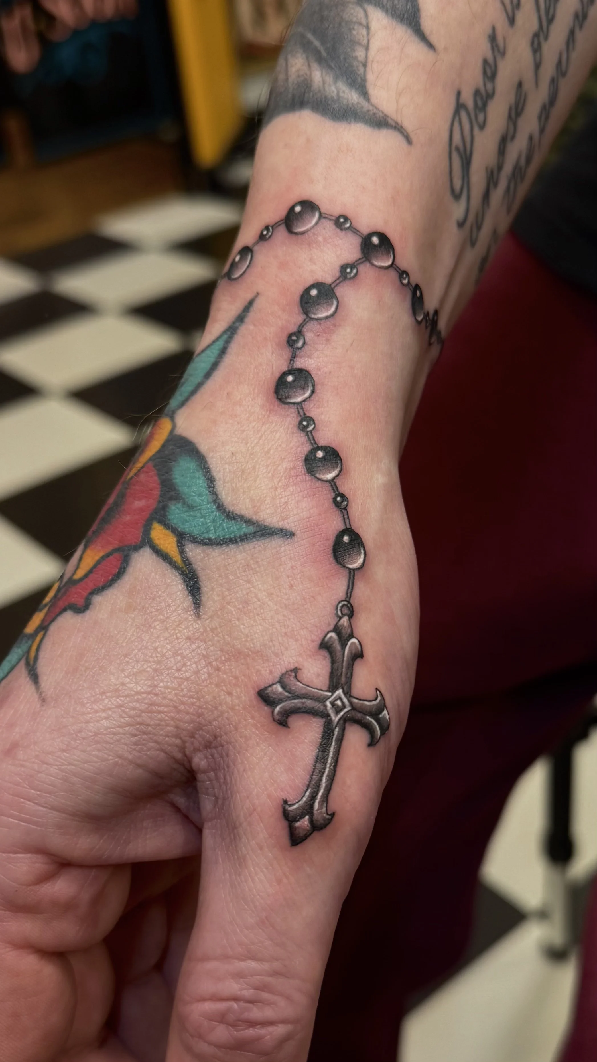 A tattoo of a rosary hanging over a tattooed hand, which features a colorful floral design.