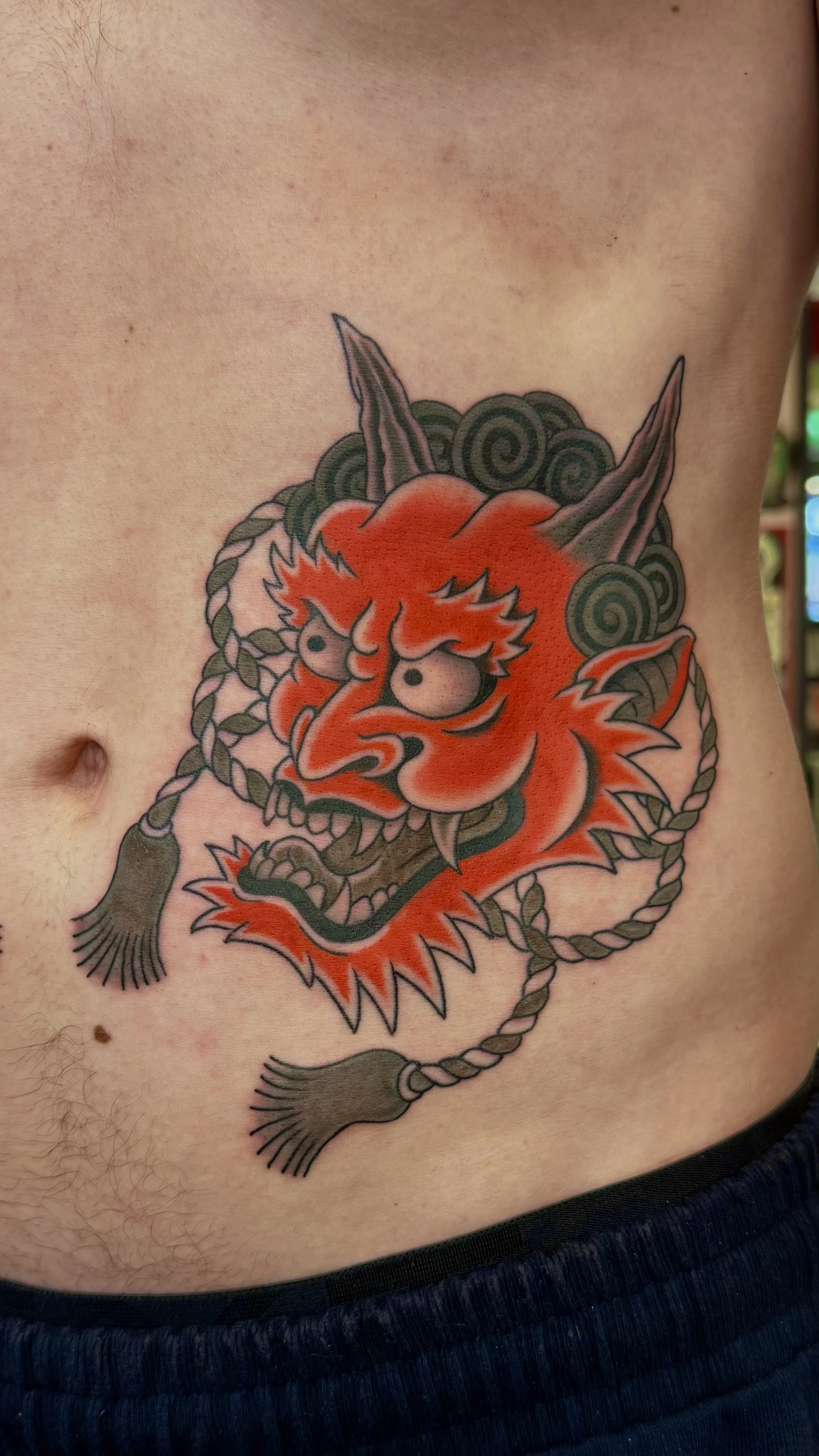 Tattoo of an angry dragon's face with orange, black, and green colors, featuring sharp teeth and whiskers, surrounded by a braided rope.