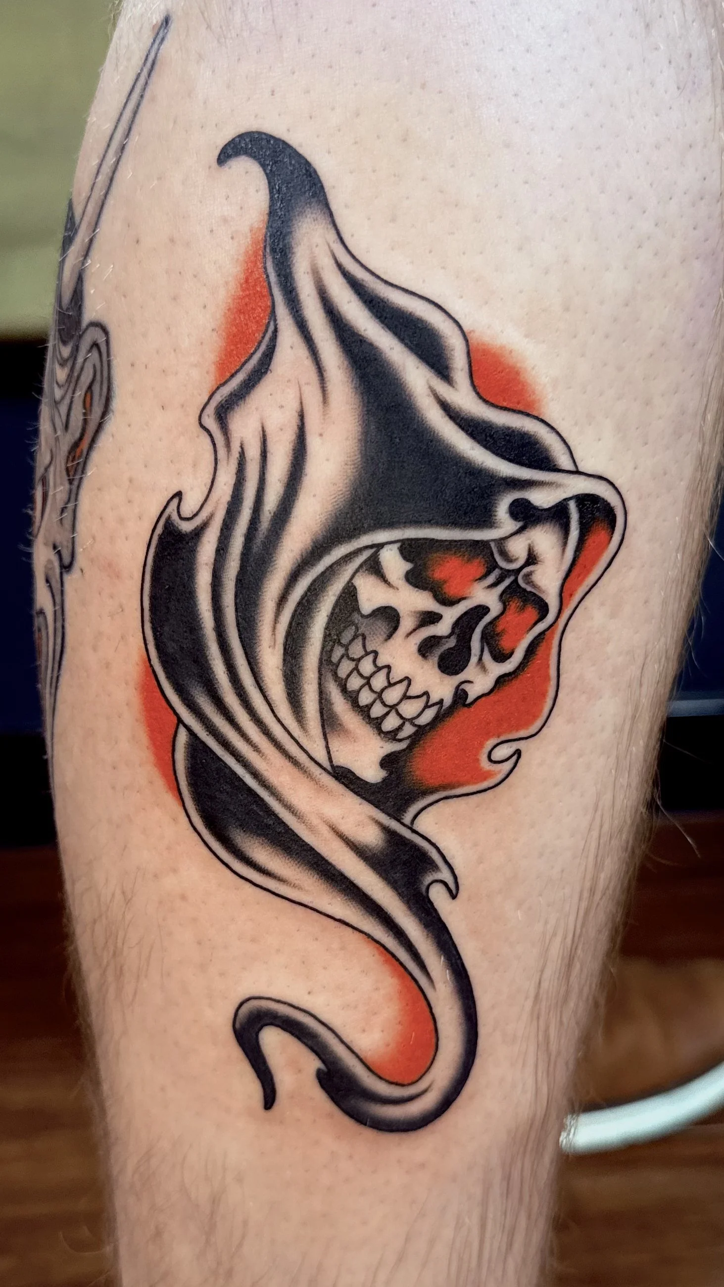 A tattoo of a skull with a hood, integrated into a stylized smoke or serpent design, with black, gray, red, and orange colors.