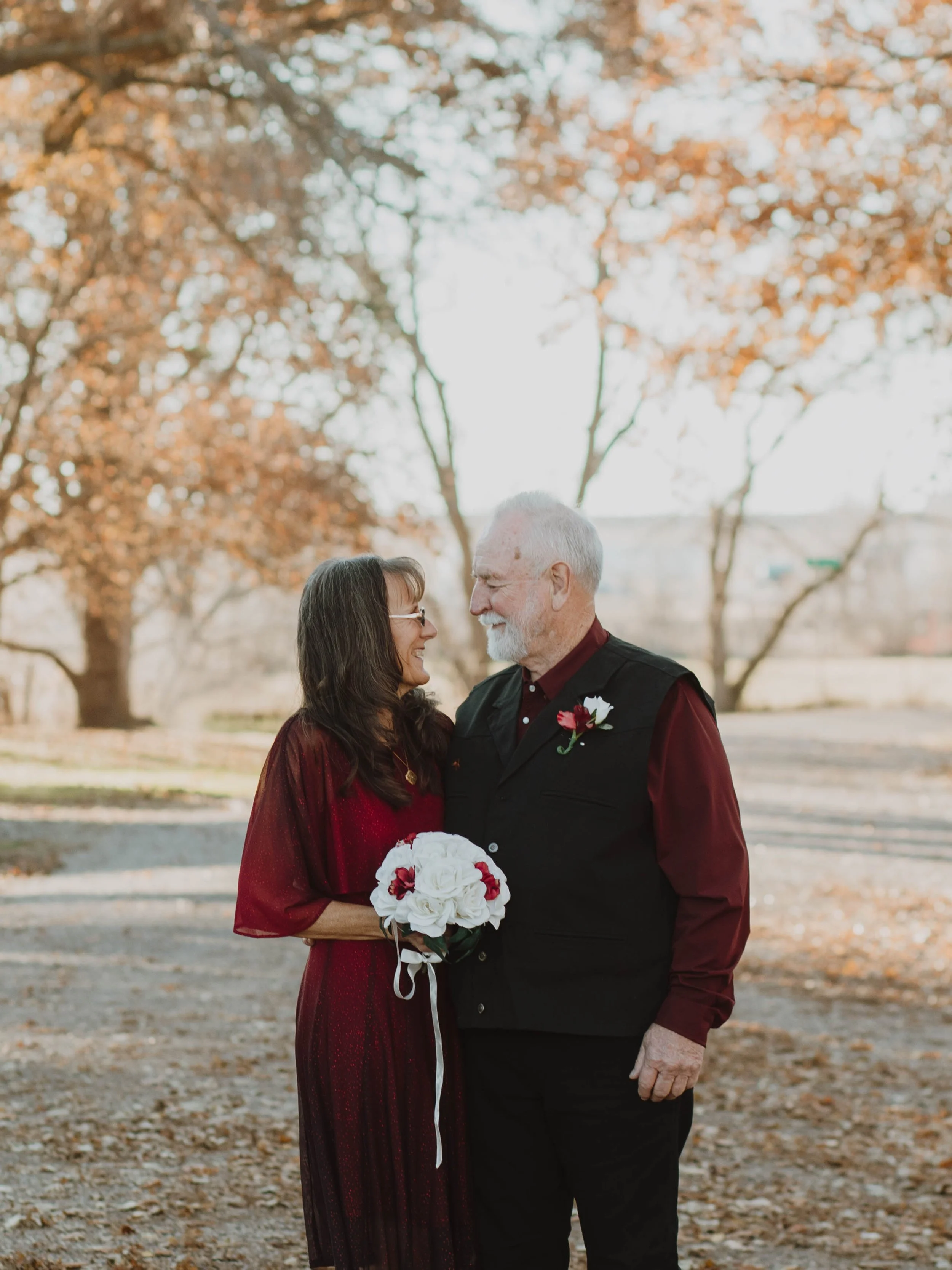 An elderly couple smiling and looking at each other outdoors during autumn, the woman is holding a bouquet of white and red flowers, and they are dressed in red and black clothing, standing on a leaf-covered path with trees in the background.