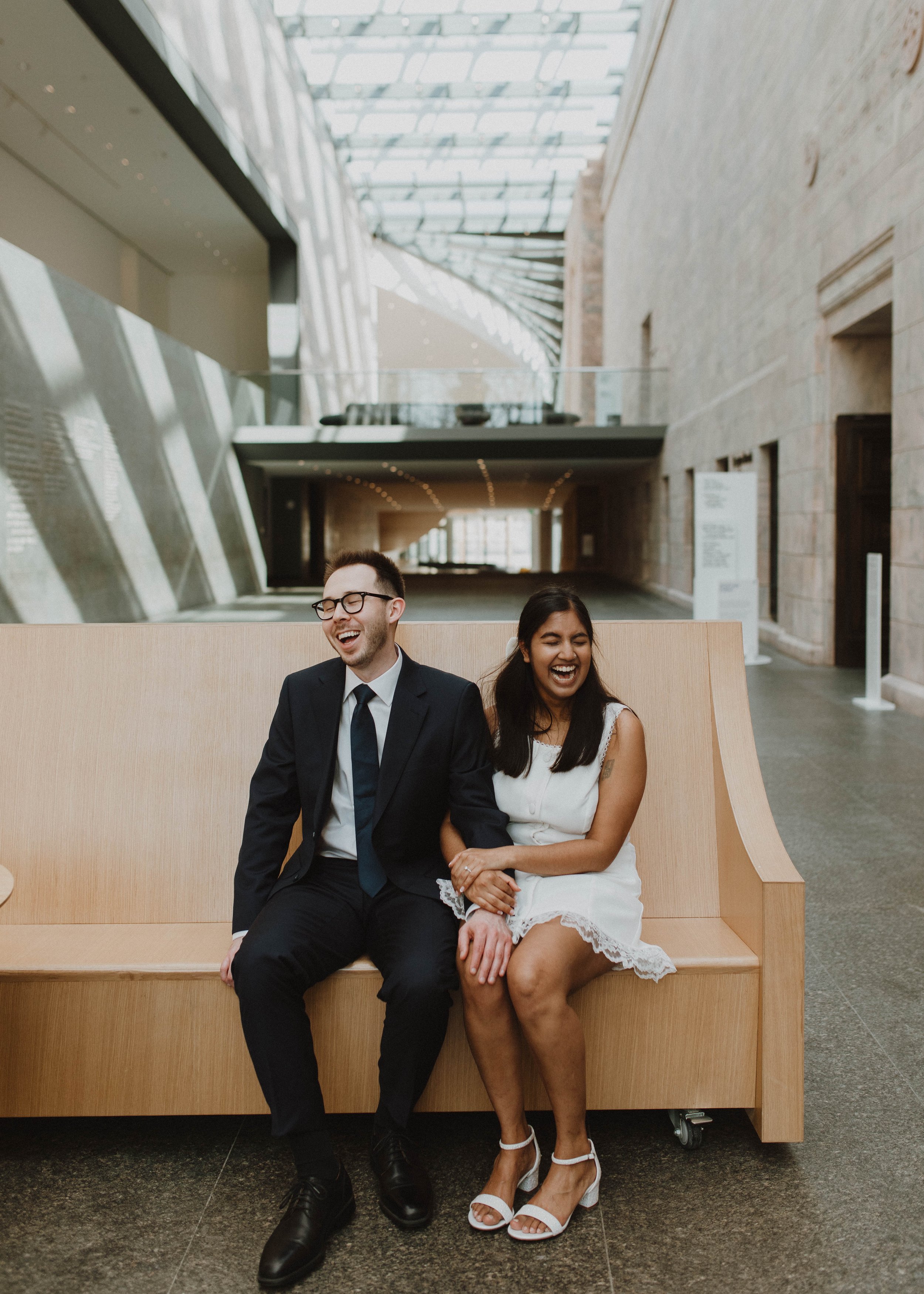 A man in a suit and a woman in a white dress laughing and sitting together on a wooden bench in the Joslyn Art Museum with a glass ceiling and stone walls.