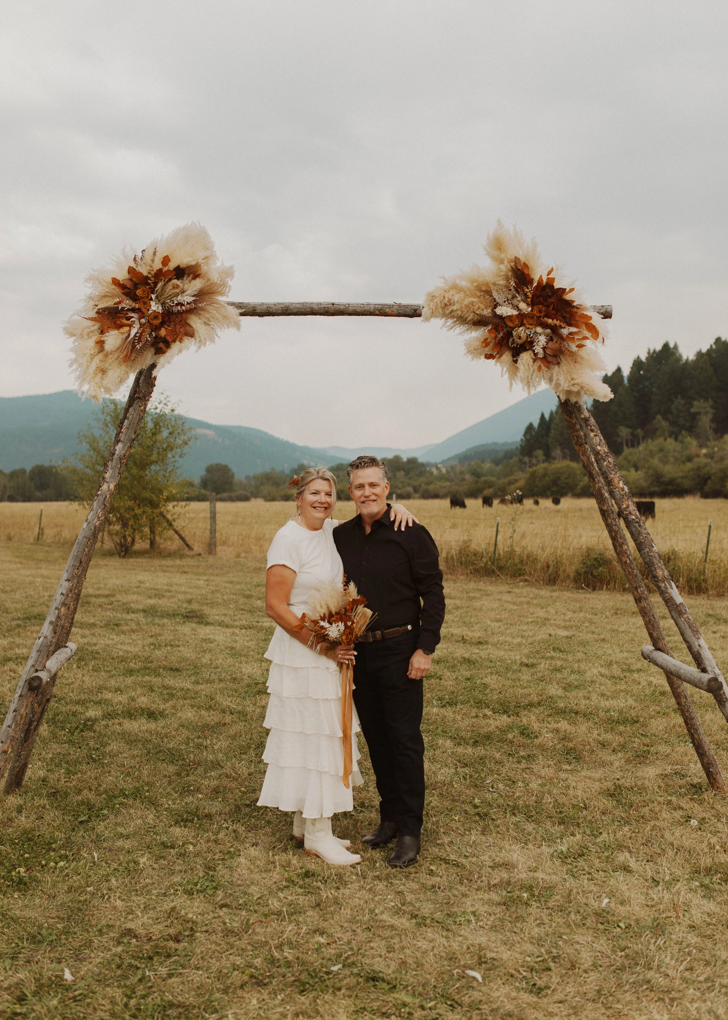 A couple standing under a wooden wedding arch decorated with dried floral arrangements in a rural outdoor setting, with mountains and open fields in the background.