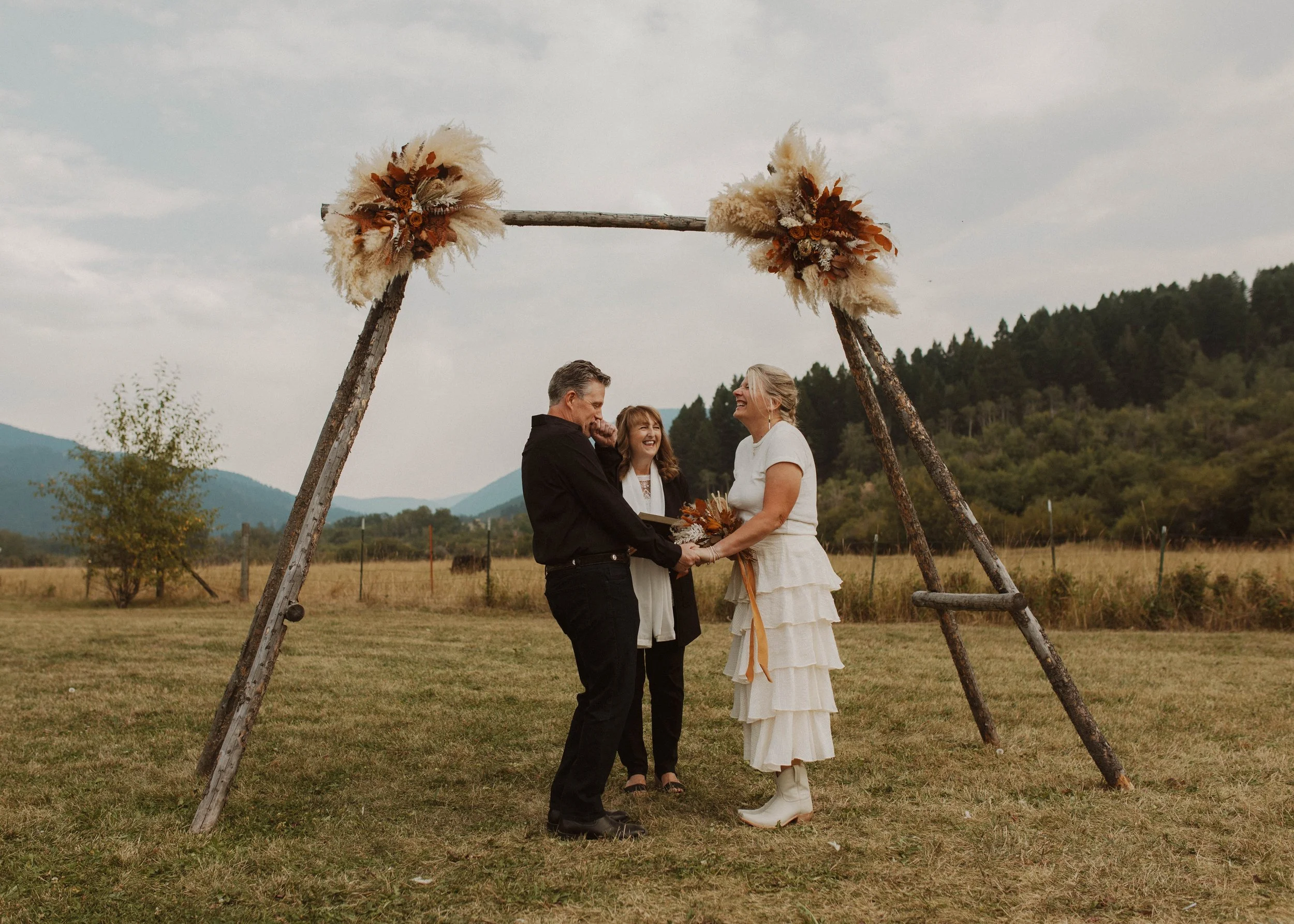 A couple getting married outdoors in Bozeman, Montana under a rustic arch decorated with dried flowers, with an officiant standing behind them, in a scenic field with mountains in the background.