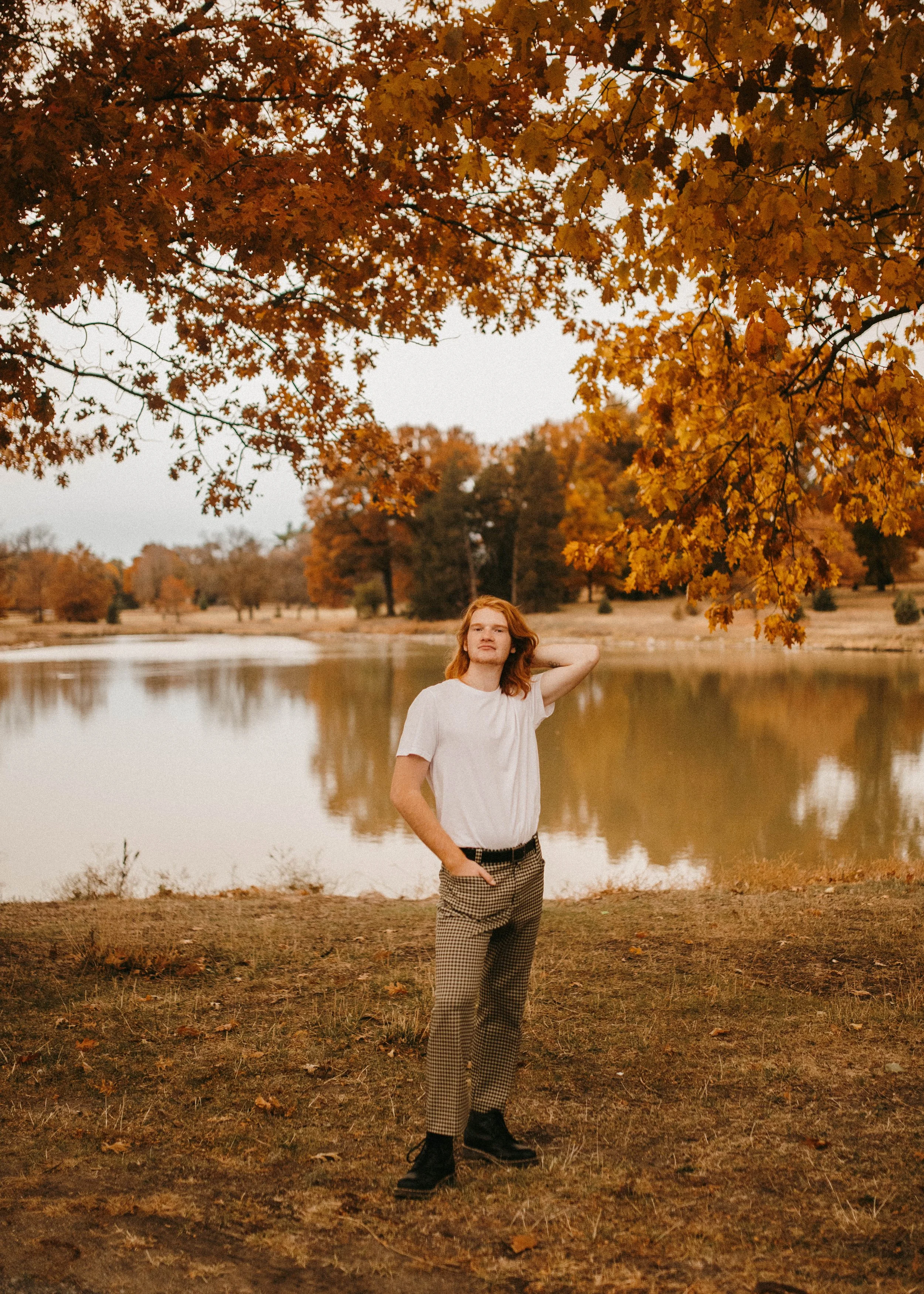 A young man with red hair standing by a lake in Pioneers Park, Lincoln, during autumn, surrounded by orange and yellow leaves.