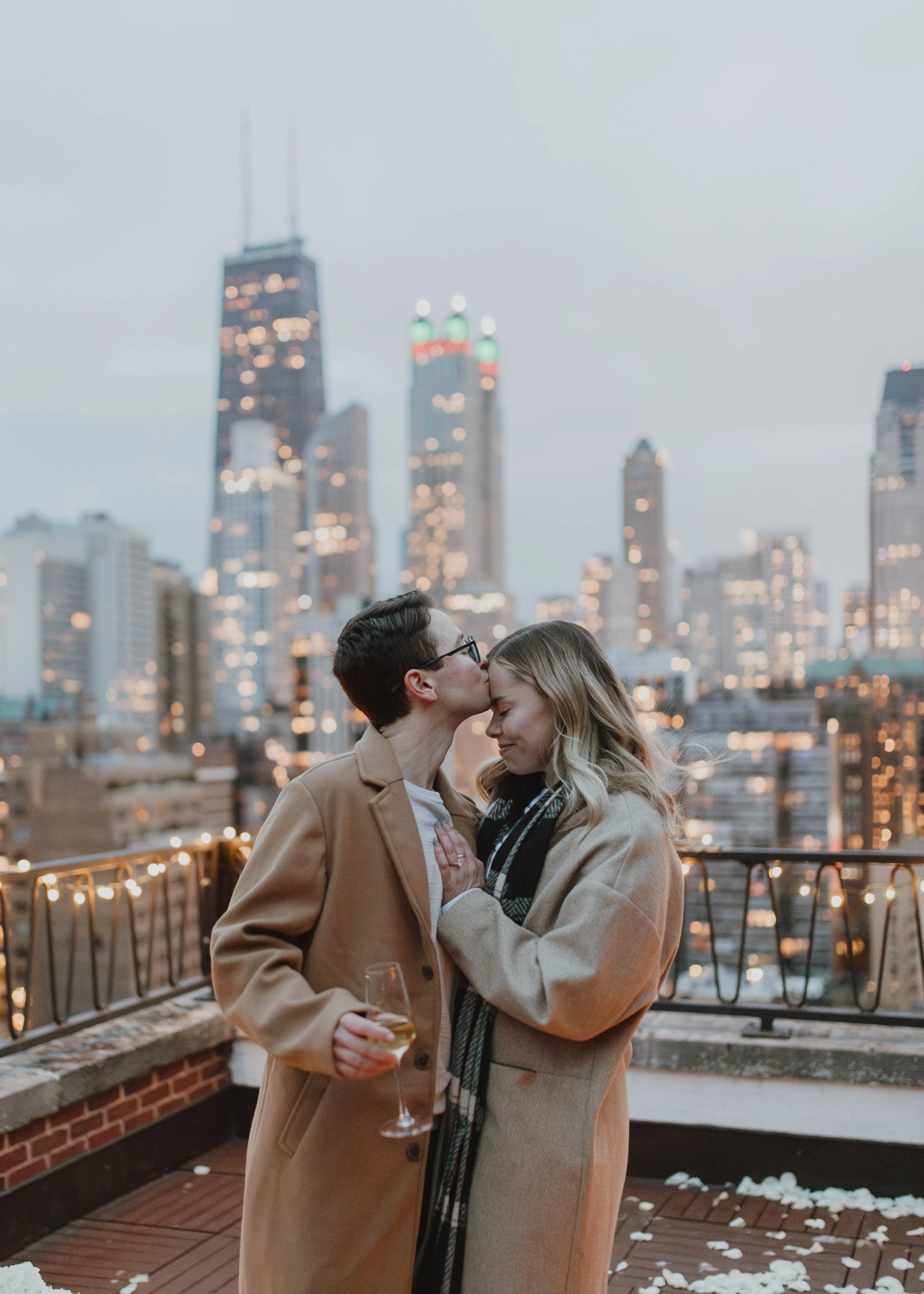 A couple on a rooftop with the Chicago skyline in the background, the man kissing the woman's forehead as they hold a glass of wine.
