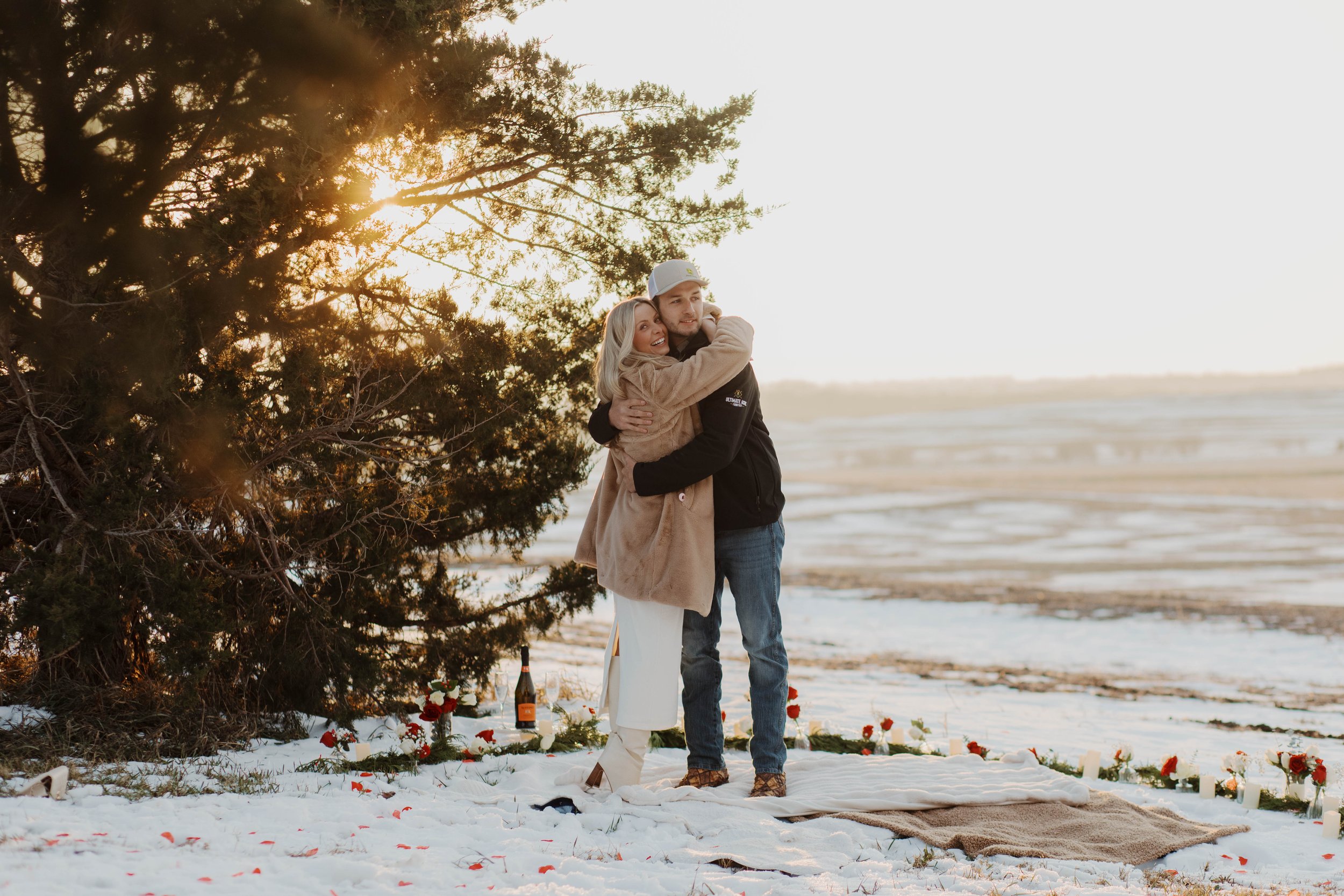 A couple hugging outdoors in Nebraska on a snow-covered patch near a large tree at sunset, with a blanket, flowers, candles, and a bottle of champagne on the ground.