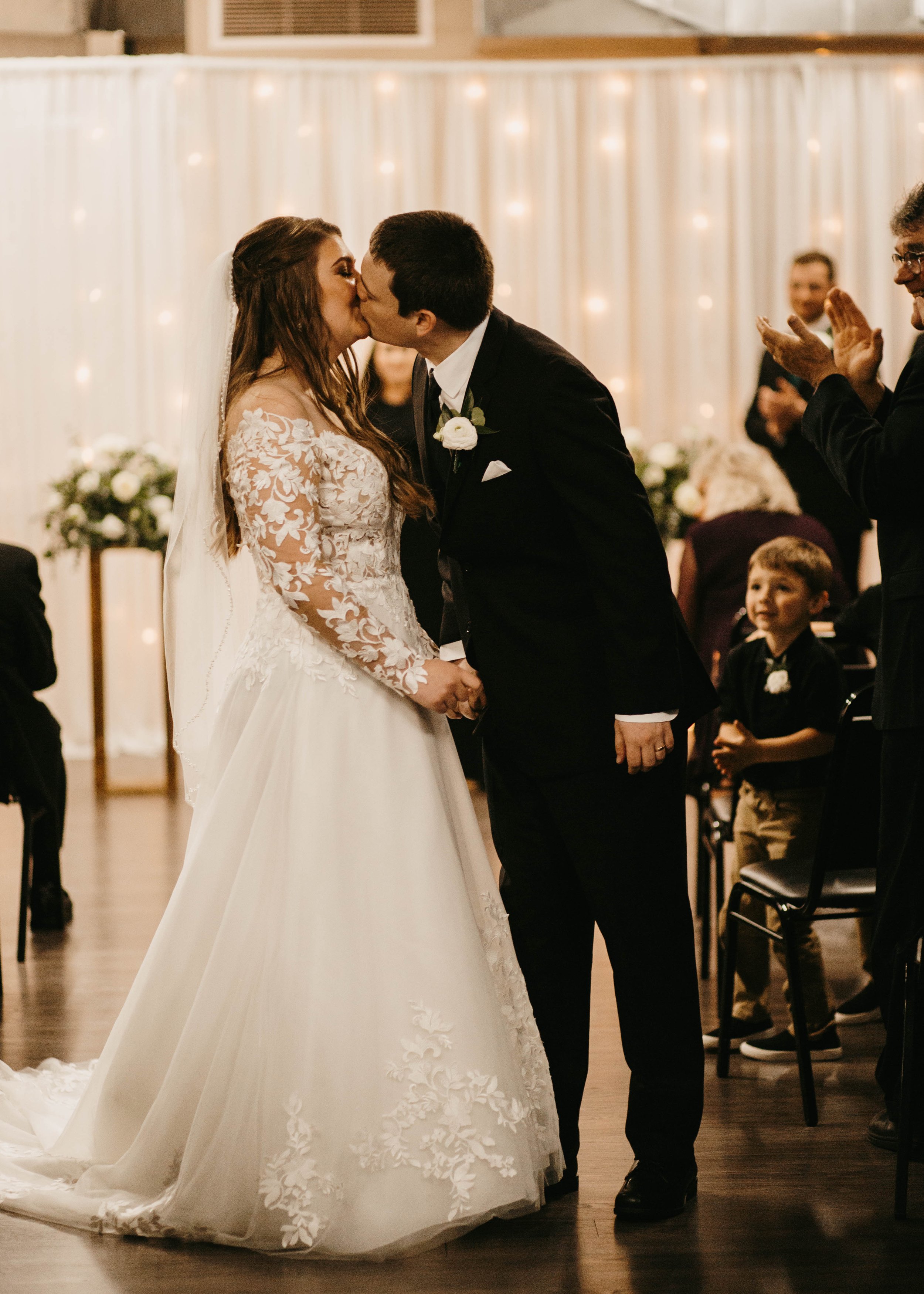 A bride and groom kiss during their wedding ceremony, holding hands, with guests clapping in the background.