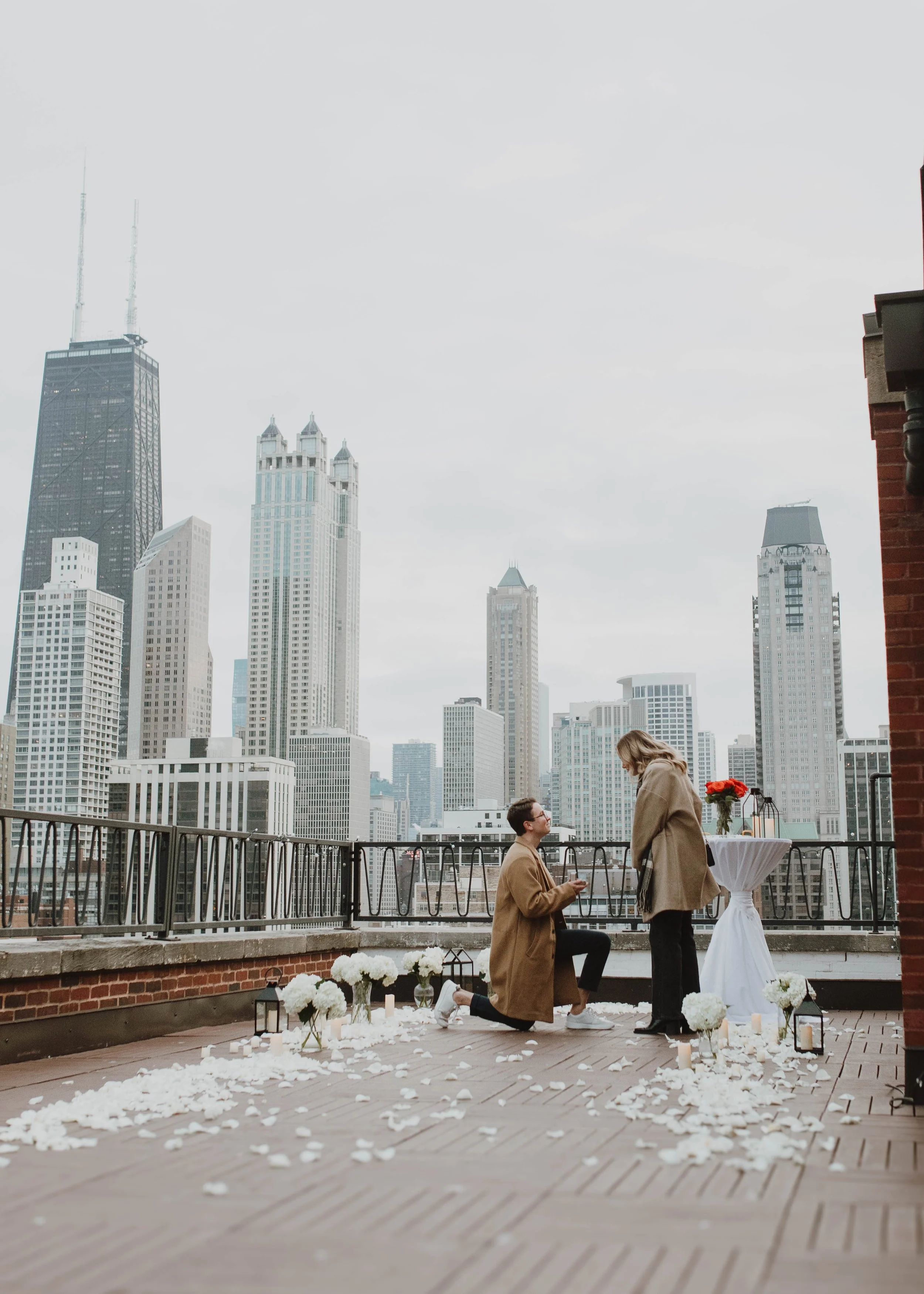 A couple on a rooftop with the Chicago skyline in the background, during a wedding proposal. The man is kneeling, proposing to the woman, with flowers, candles, and rose petals on the wooden deck.