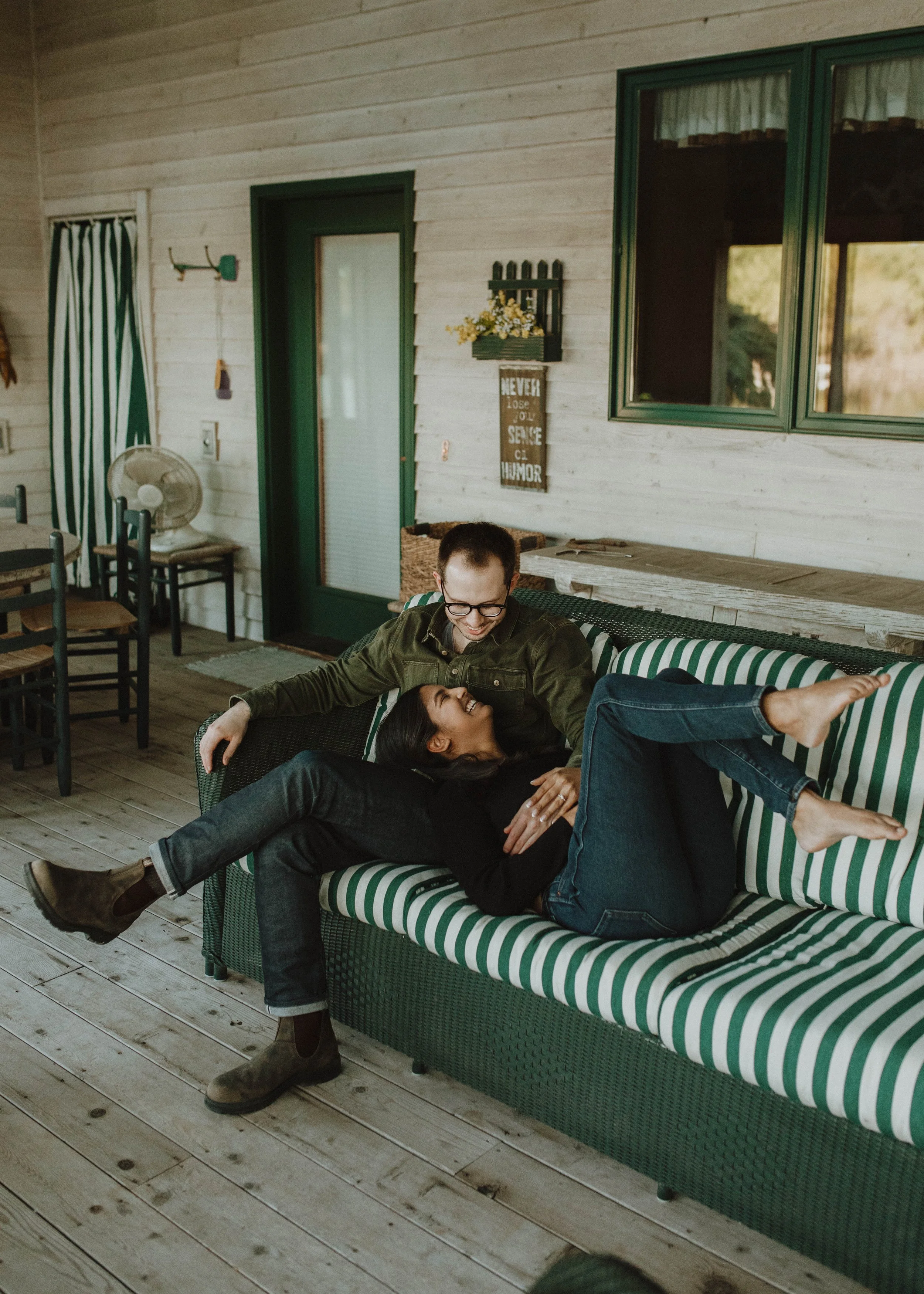 A man and woman are playfully lying on a green and white striped outdoor sofa on a wooden porch, smiling and looking at each other.