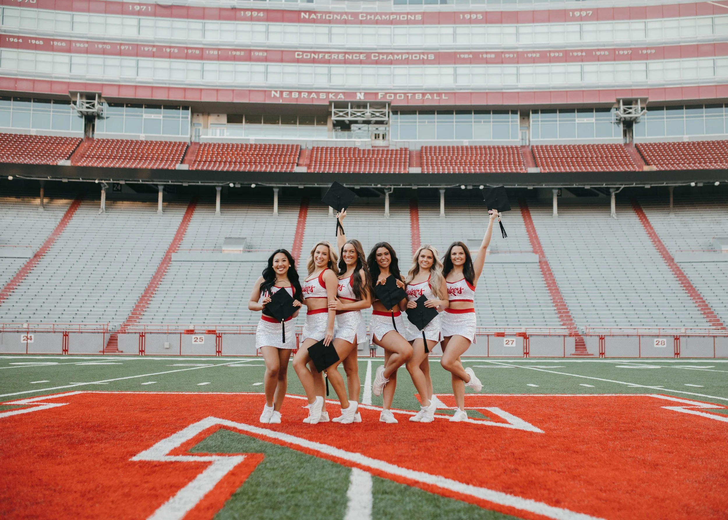 Group of six female University of Nebraska students celebrating in Memorial Stadium, wearing dance team uniforms and holding graduation caps in the air.