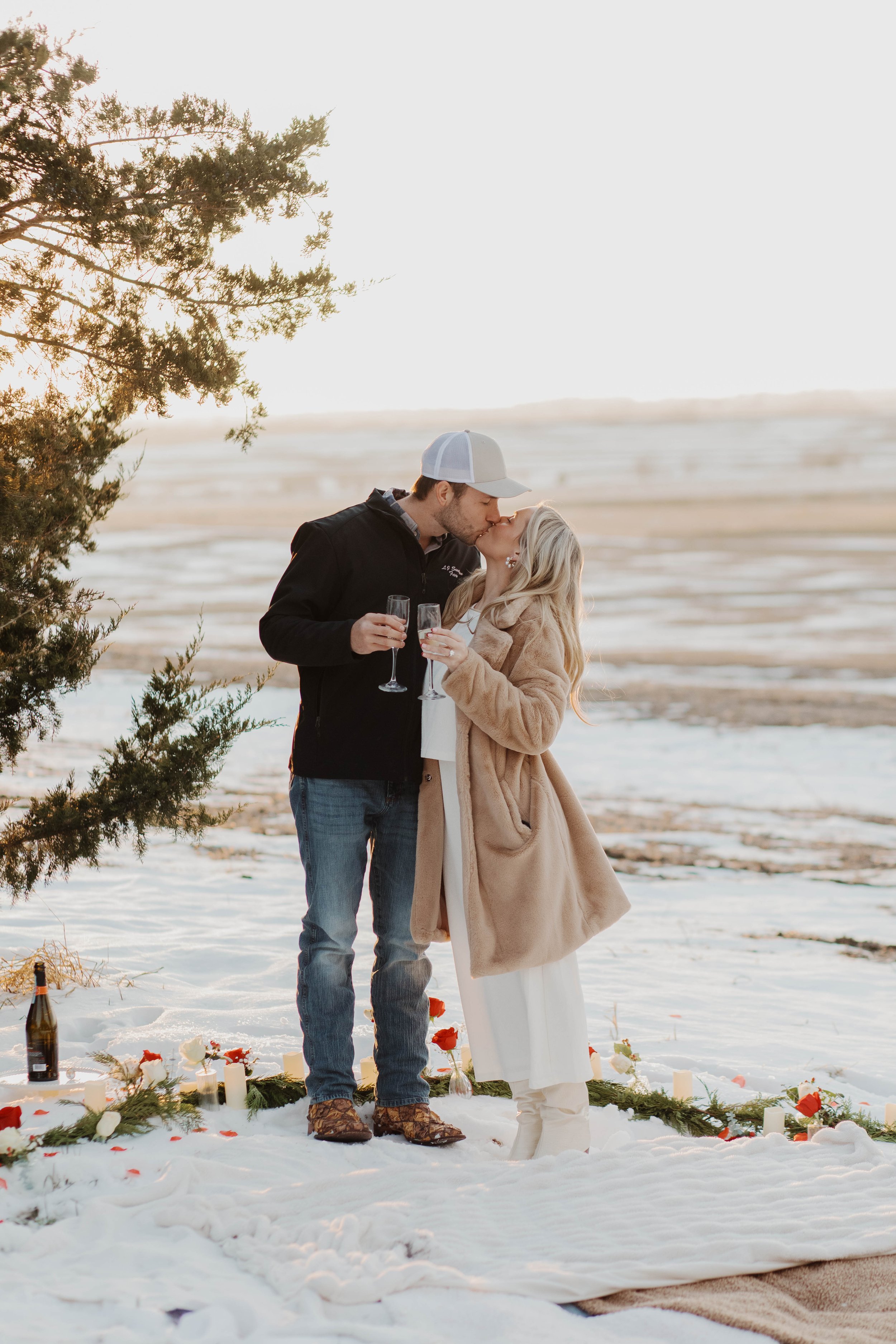 A couple kissing during a winter proposal on a snowy field in Nebraska, holding champagne glasses, with candles and flowers on the snow-covered ground.