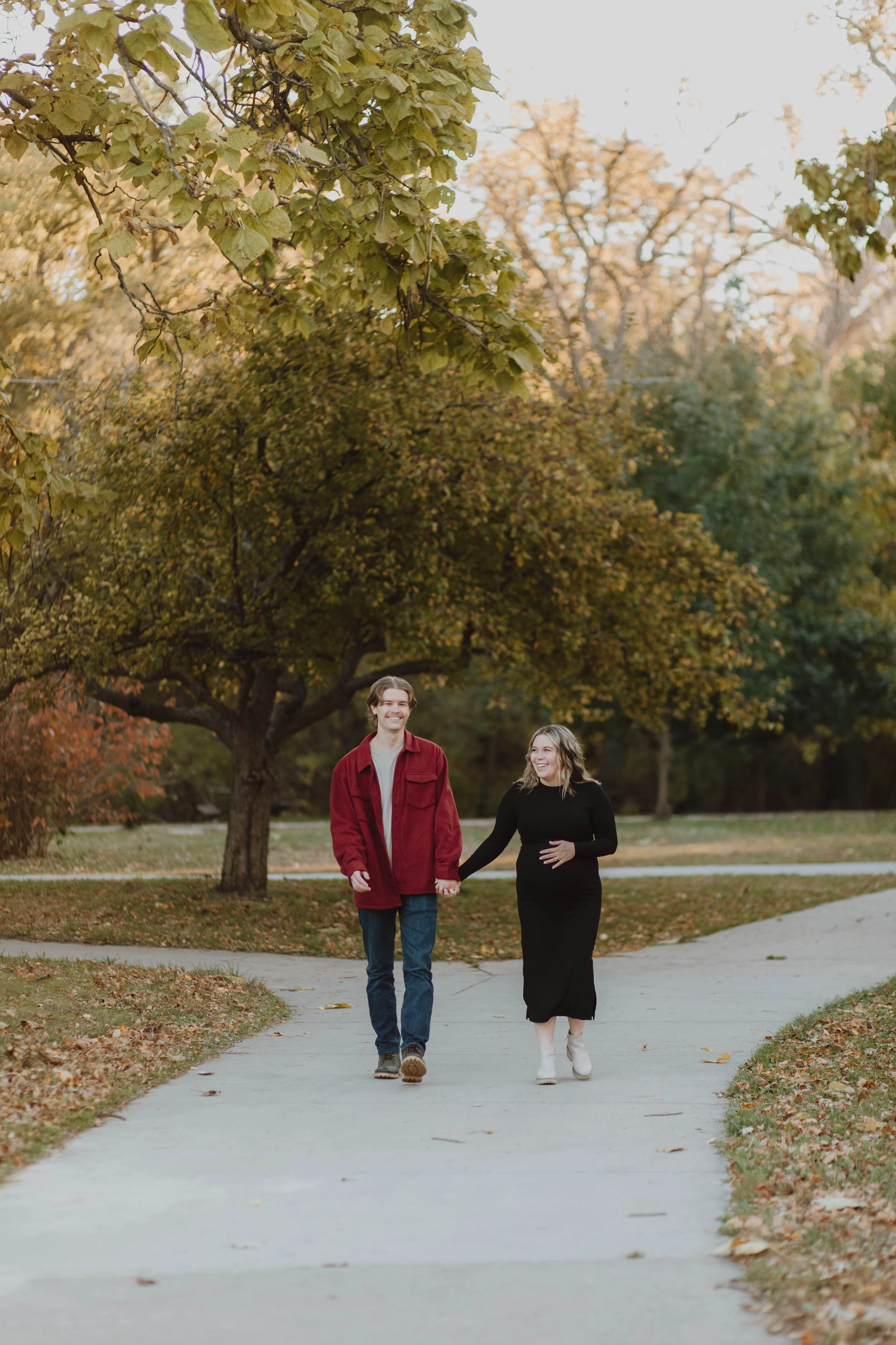 A couple walking hand in hand on a paved path in Elmwood Park, Omaha during autumn, with colorful fall foliage on trees.