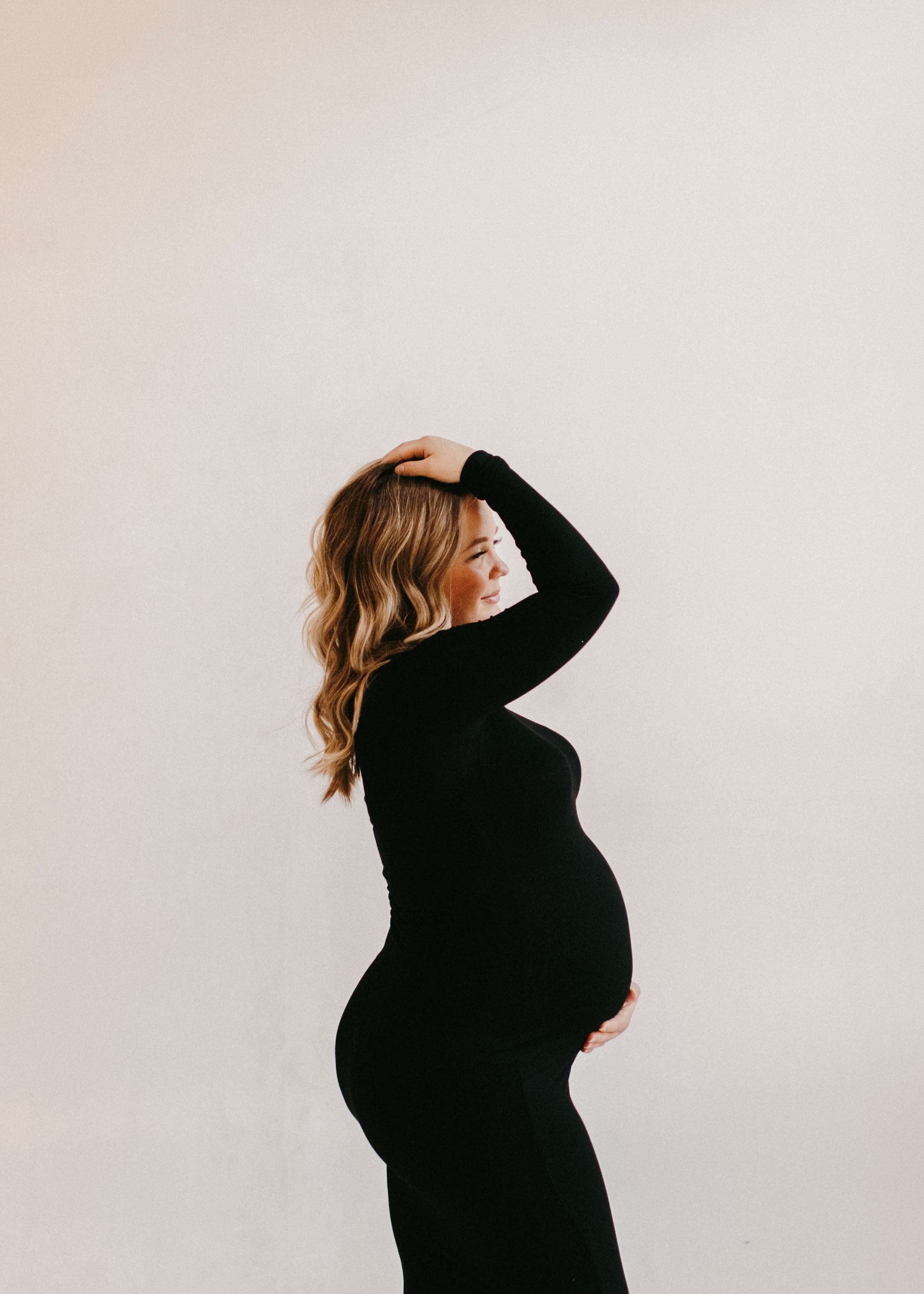 A pregnant woman in a black long-sleeve outfit, standing sideways with her eyes closed, touching her head with one hand and holding her belly with the other, against a plain white background.