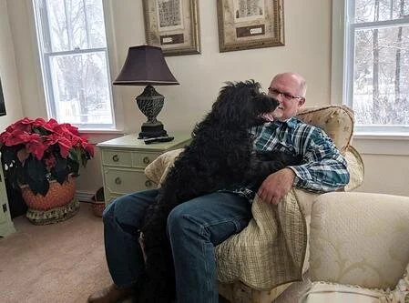 A man sitting on a beige armchair with a black dog on his lap, inside a well-lit living room with windows, a nightstand with a lamp, and a poinsettia plant.