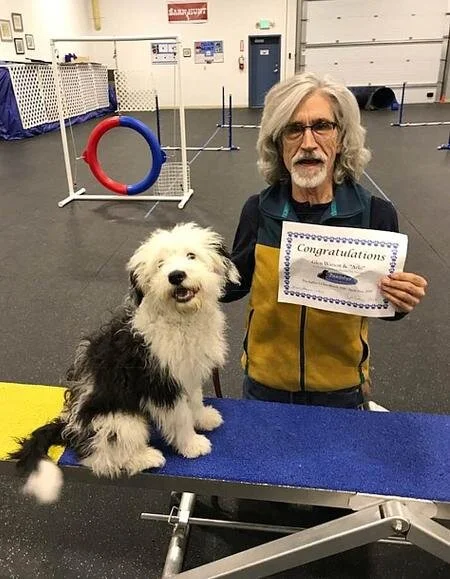 A man holding a certificate of congratulations next to a fluffy dog in a training or agility facility.